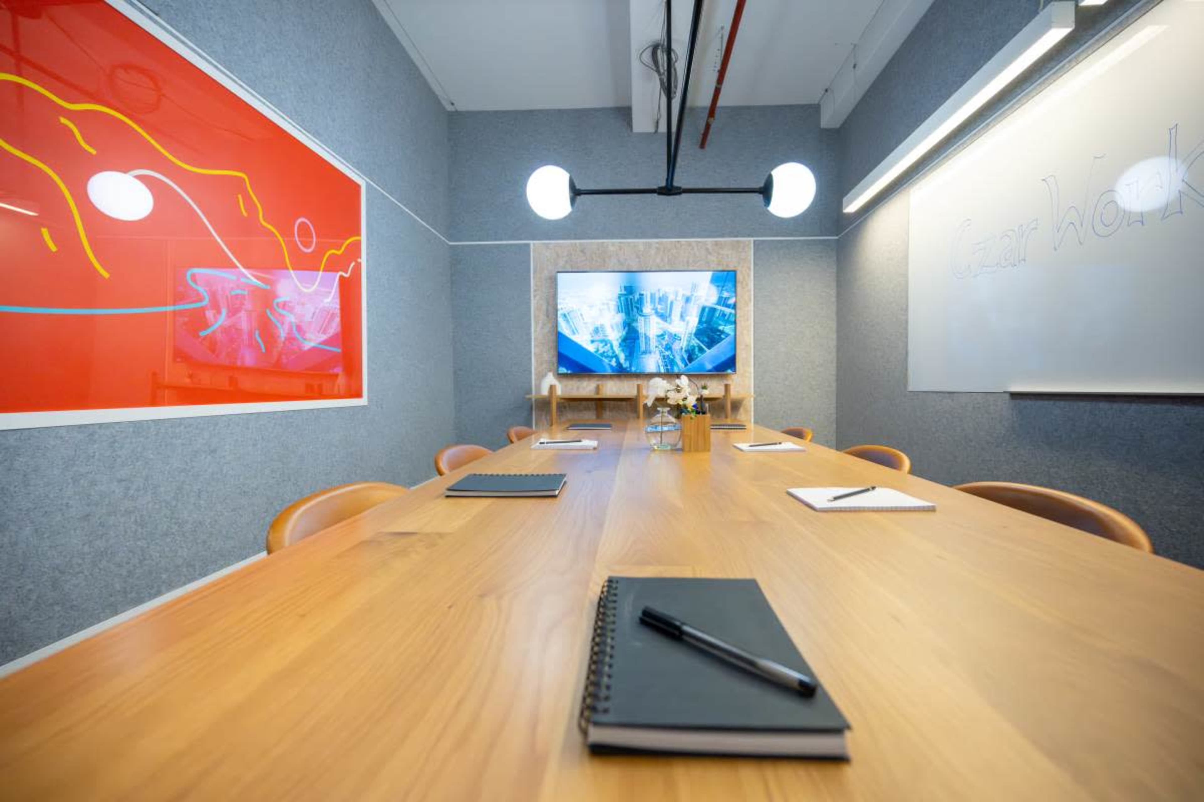A modern conference room features a long wooden table with notebooks and pens, a large screen displaying visuals, and walls adorned with abstract artwork.