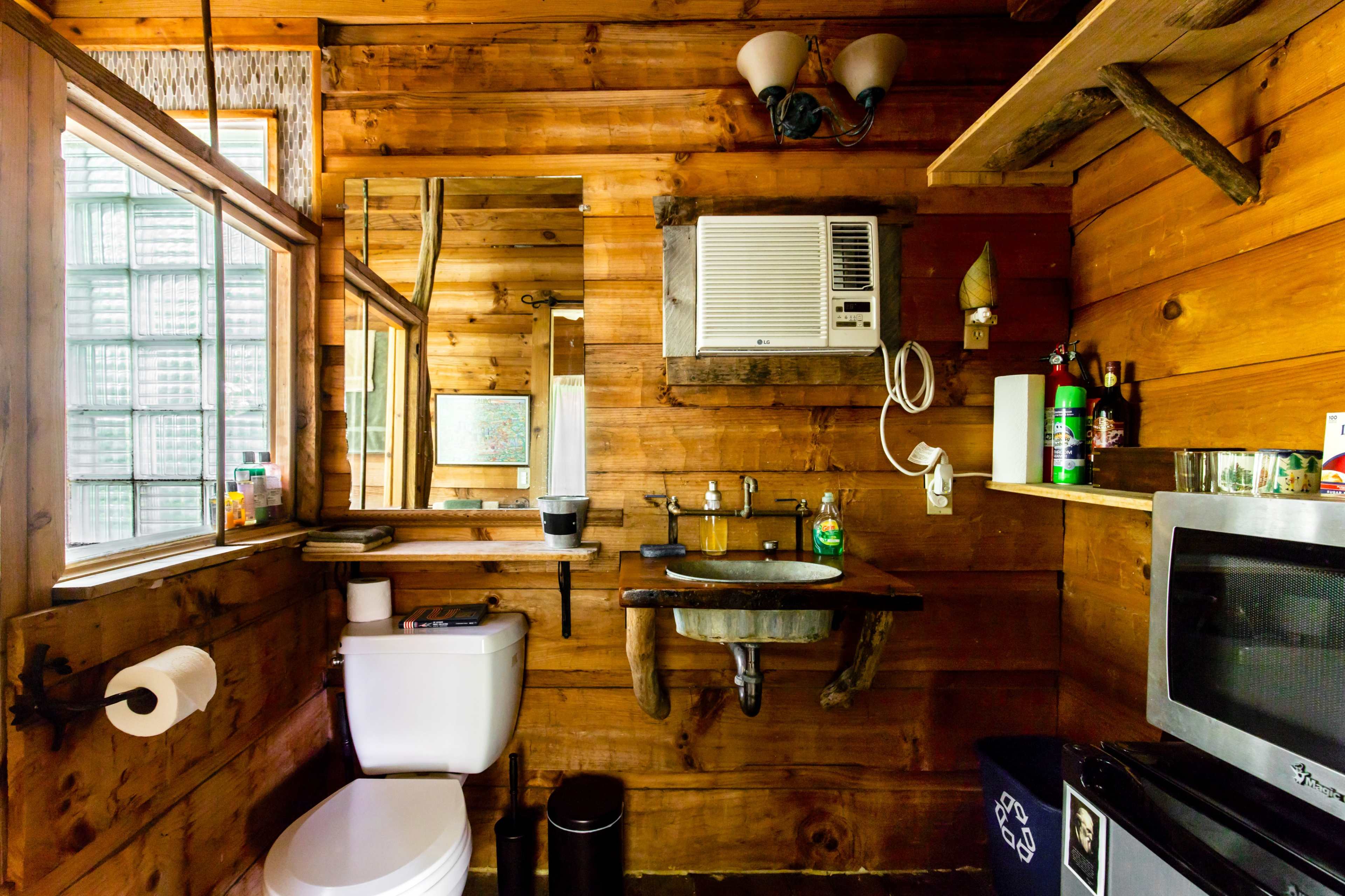 A rustic bathroom features wooden walls, a toilet, a wall-mounted sink, an air conditioning unit, and a small window.