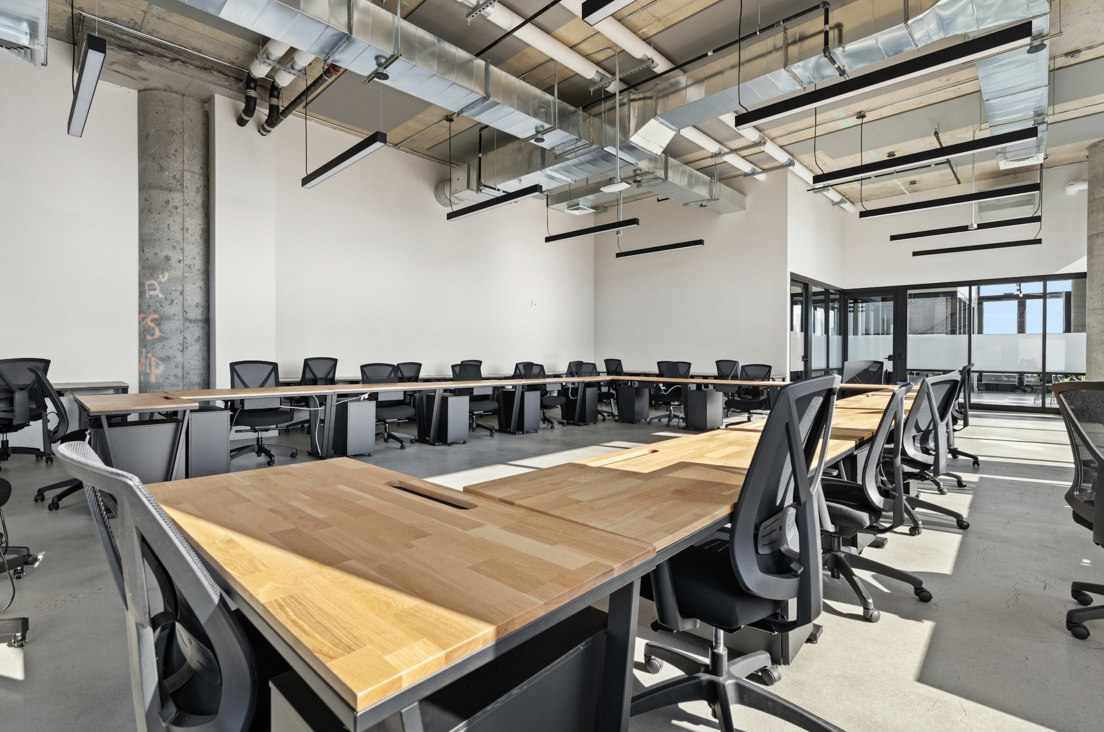 The image shows a modern, spacious office with multiple wooden tables arranged in a U-shape and black ergonomic chairs facing the center.