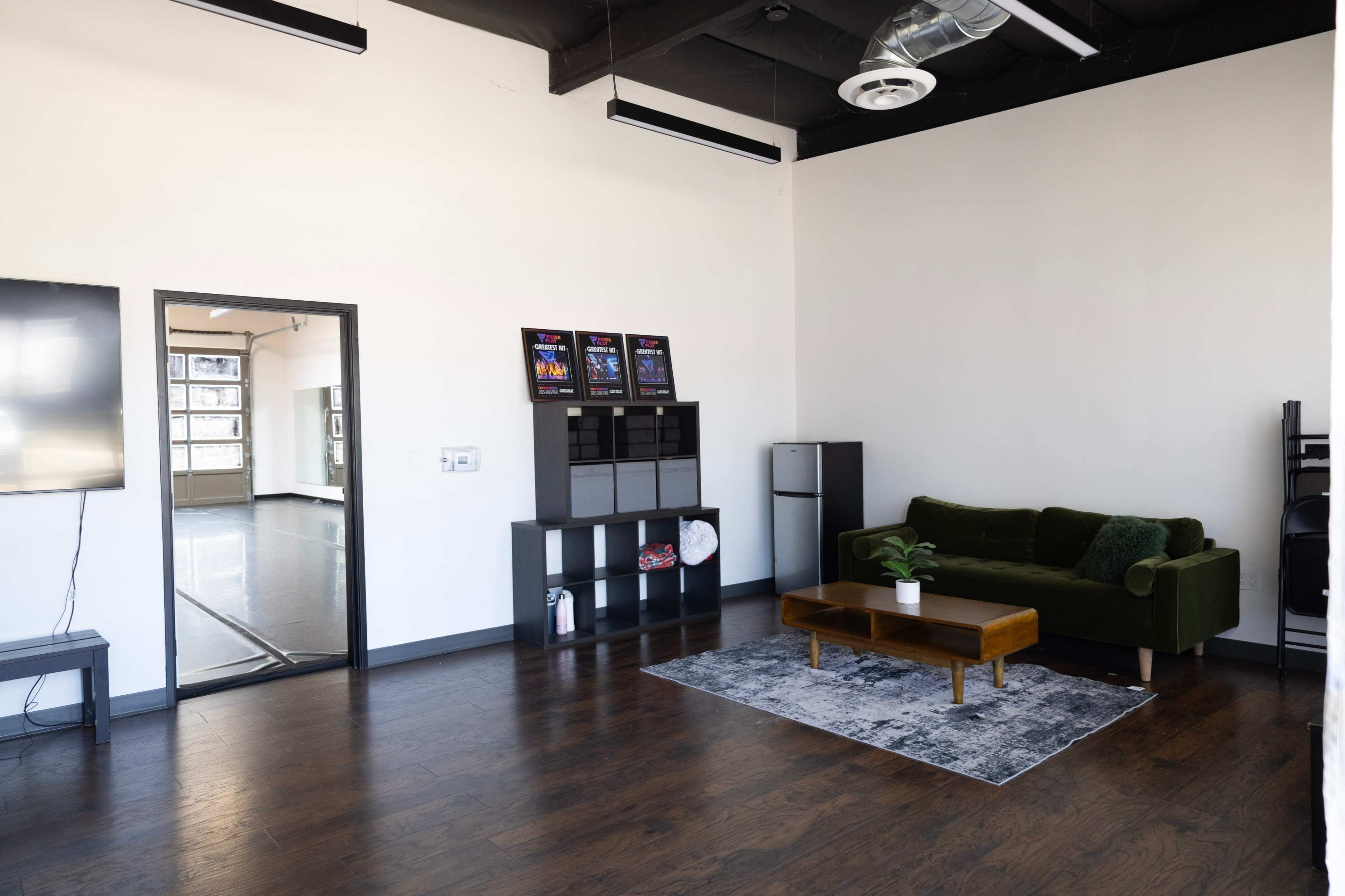 The image shows a spacious room with hardwood floors featuring a green couch, a coffee table, and a small refrigerator, with a mirror doorway leading to an adjacent area.
