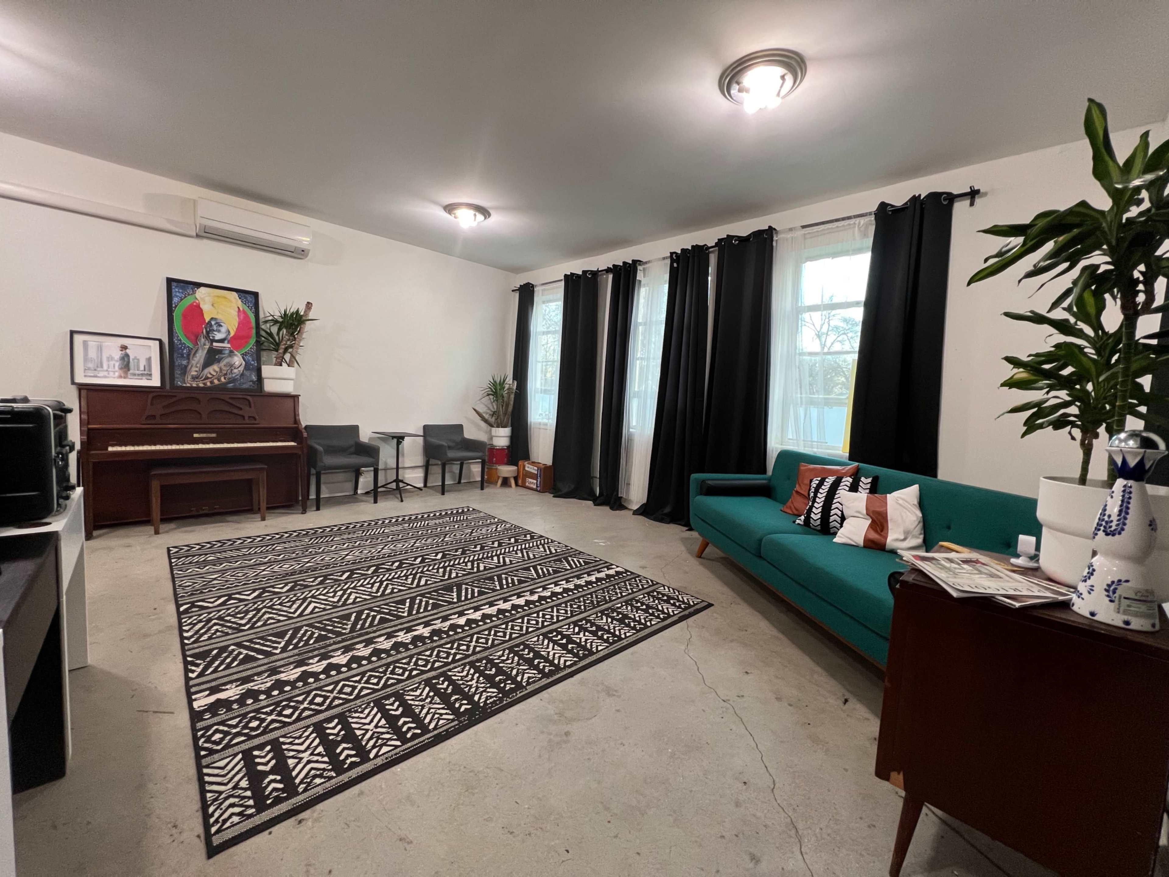 A spacious indoor living area featuring a piano, a green couch, a black and white patterned rug, and large windows with dark curtains.