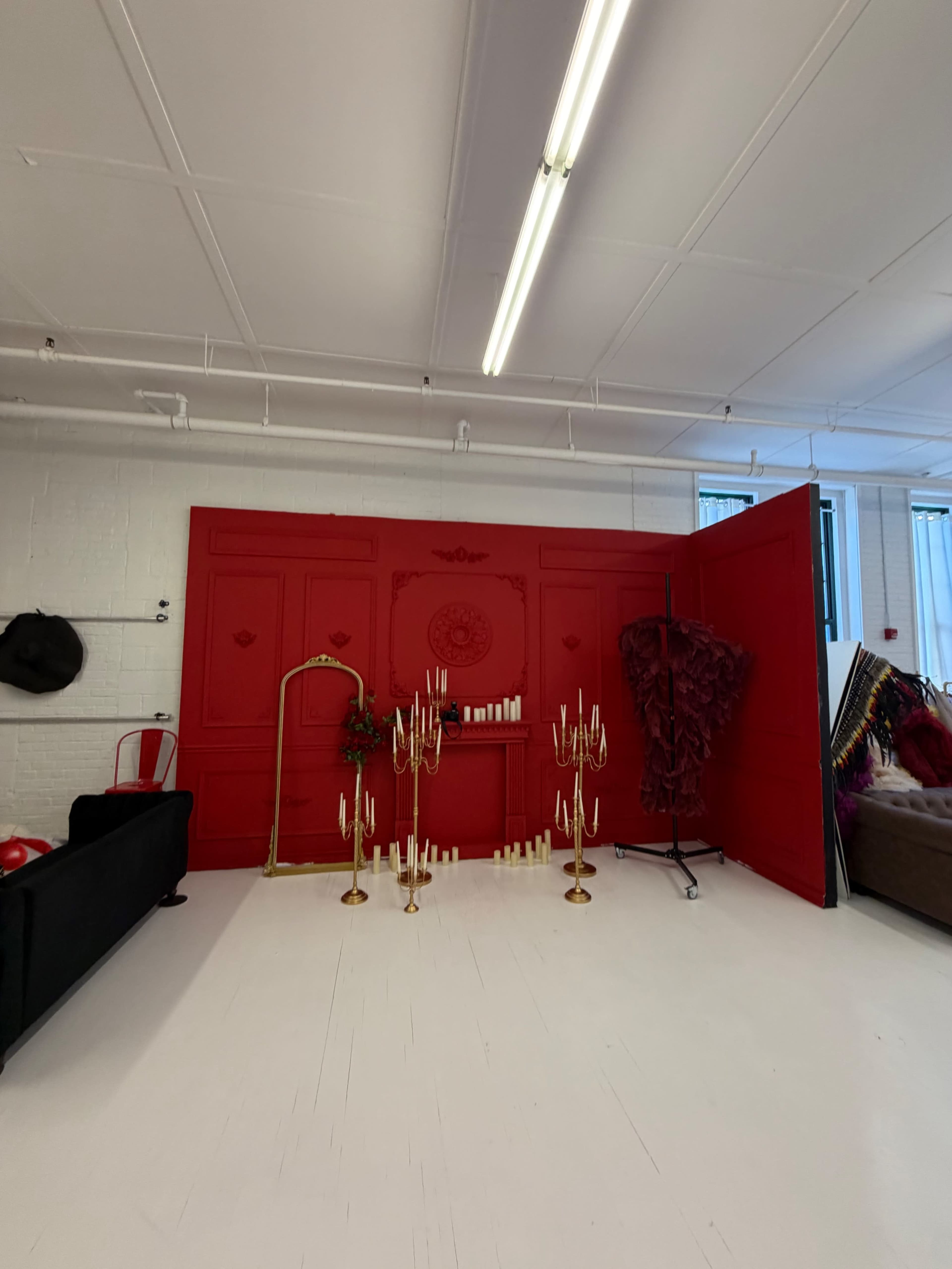 The scene features a bright red wall with decorative elements and a variety of props, including gold candelabras and a textured fabric installation, set in a spacious room with white flooring.