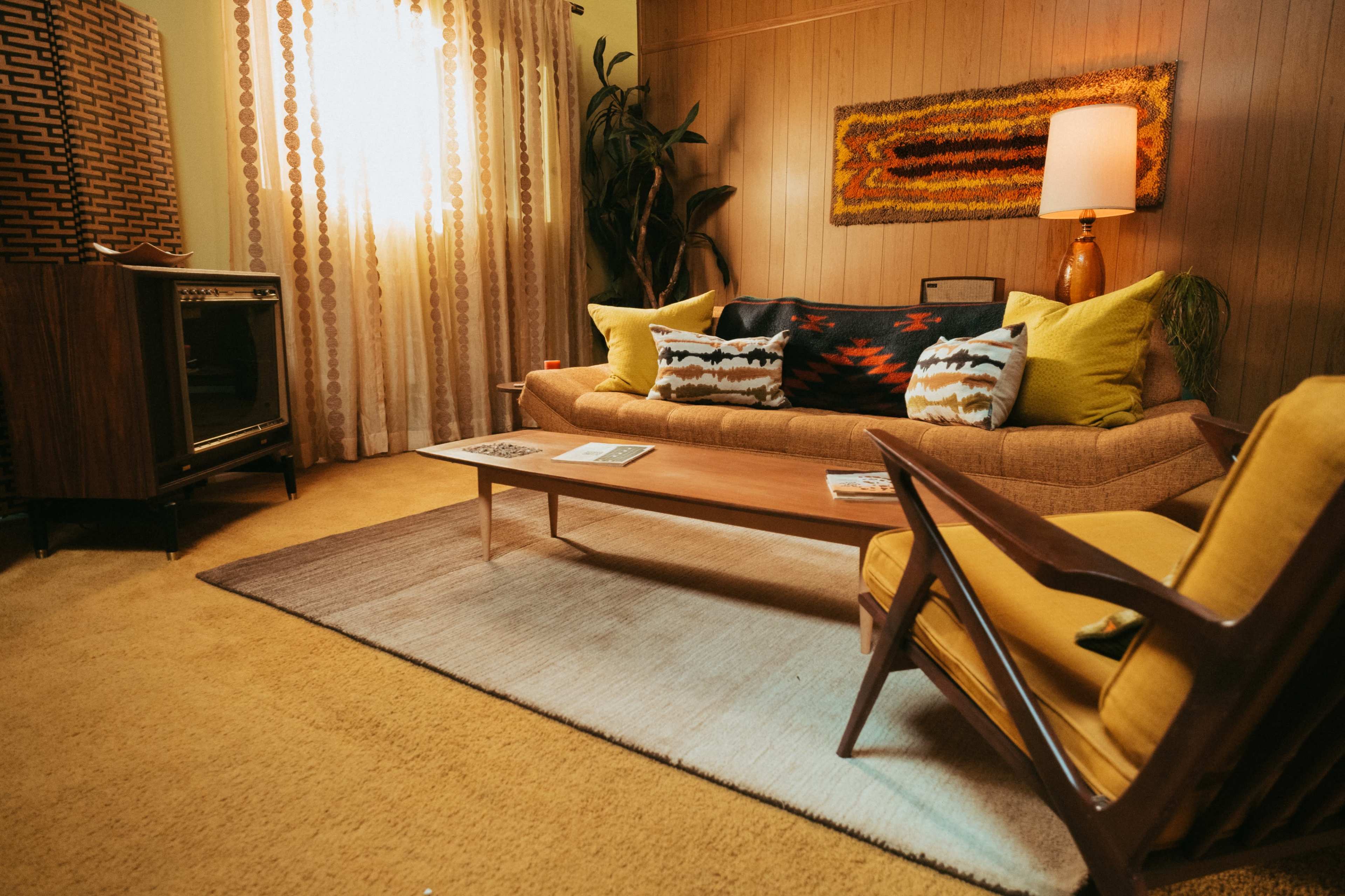 The image shows a vintage living room featuring a sofa with colorful cushions, a wooden coffee table, and a retro television set beside a large window with light curtains.
