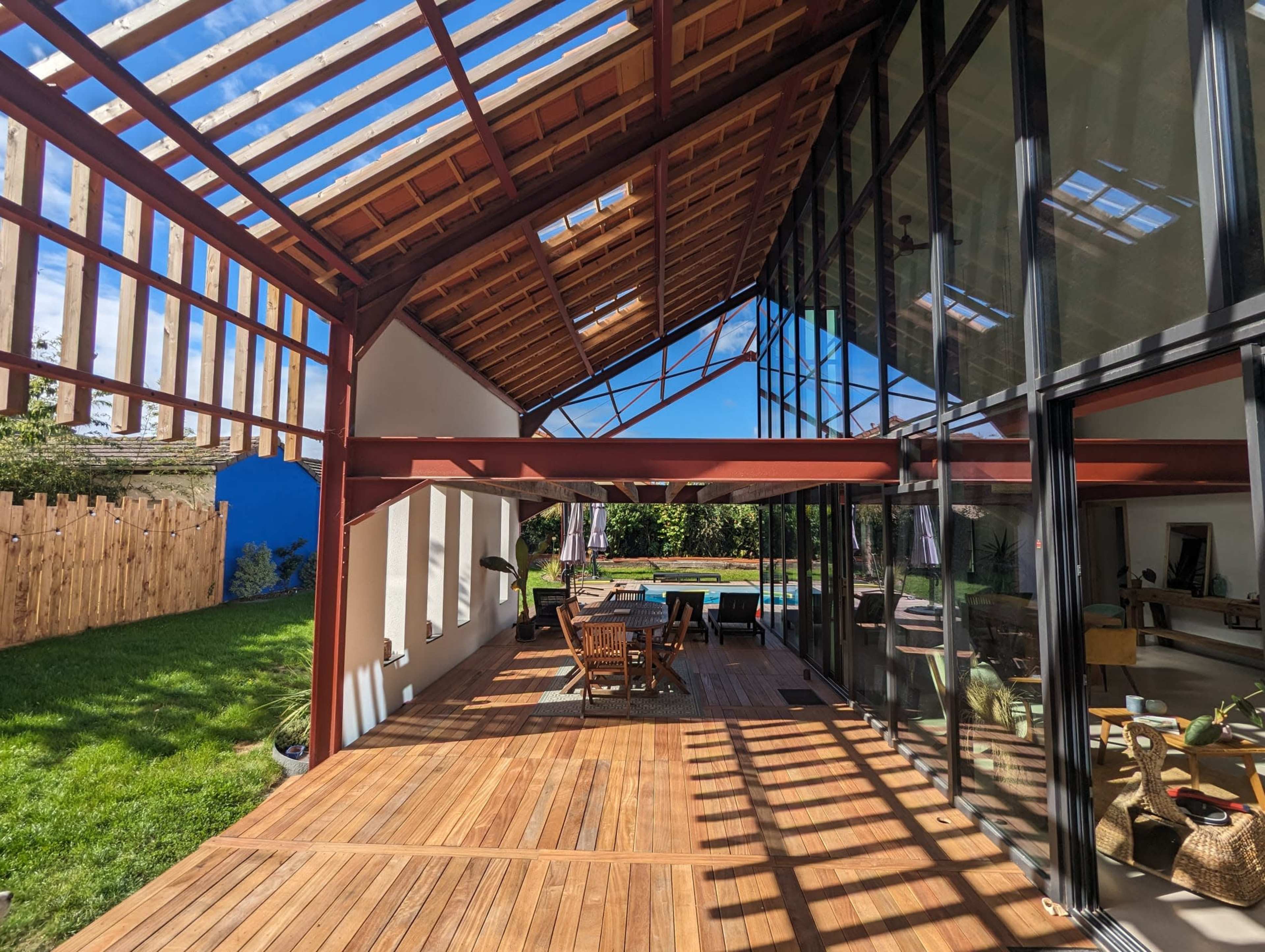 A modern outdoor patio area of a house with large glass walls, wooden decking, and a view of a green lawn and pool.