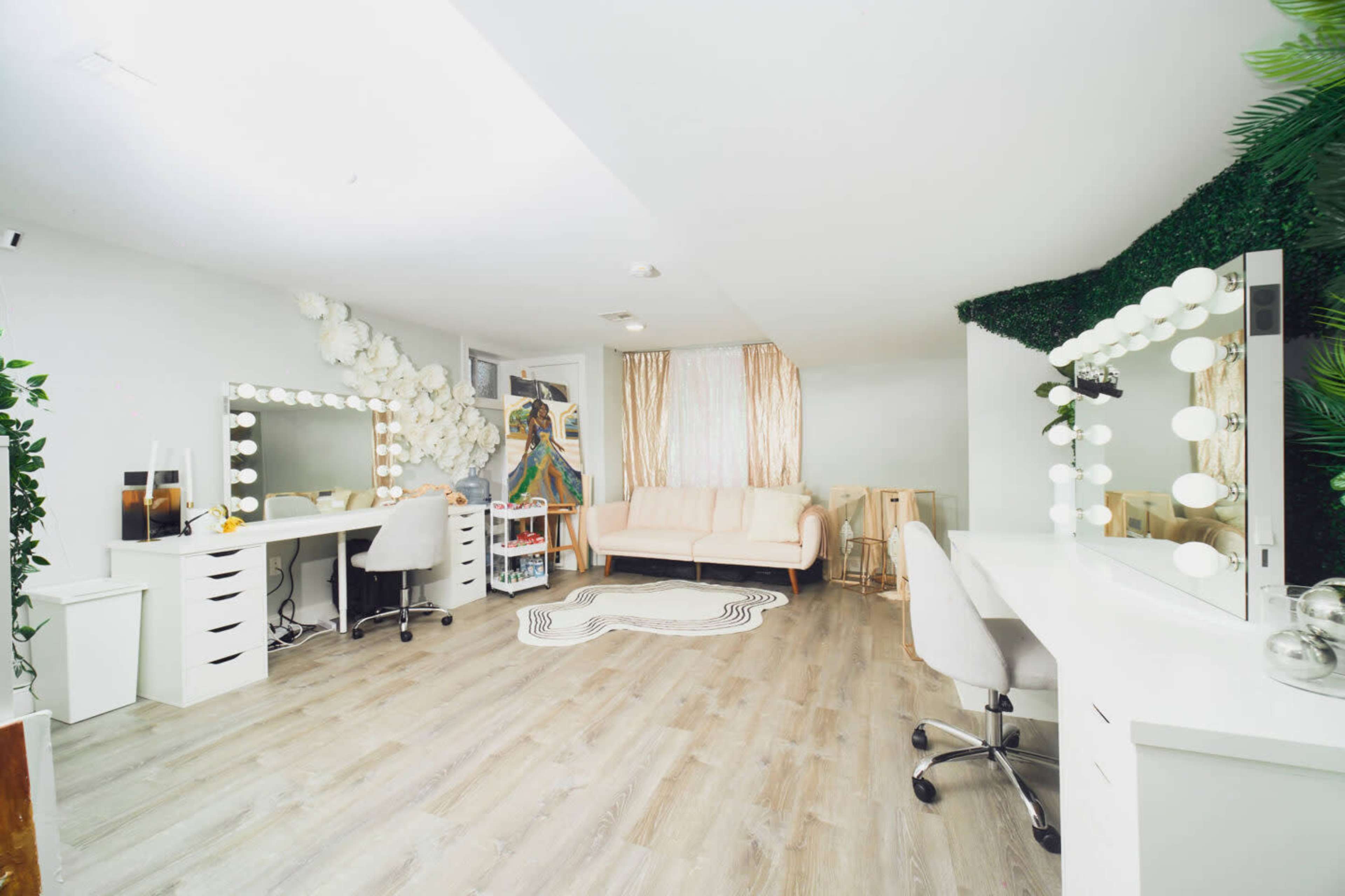 A modern, well-lit beauty studio with two large vanity stations, a cozy sofa, and decorative greenery on one wall.