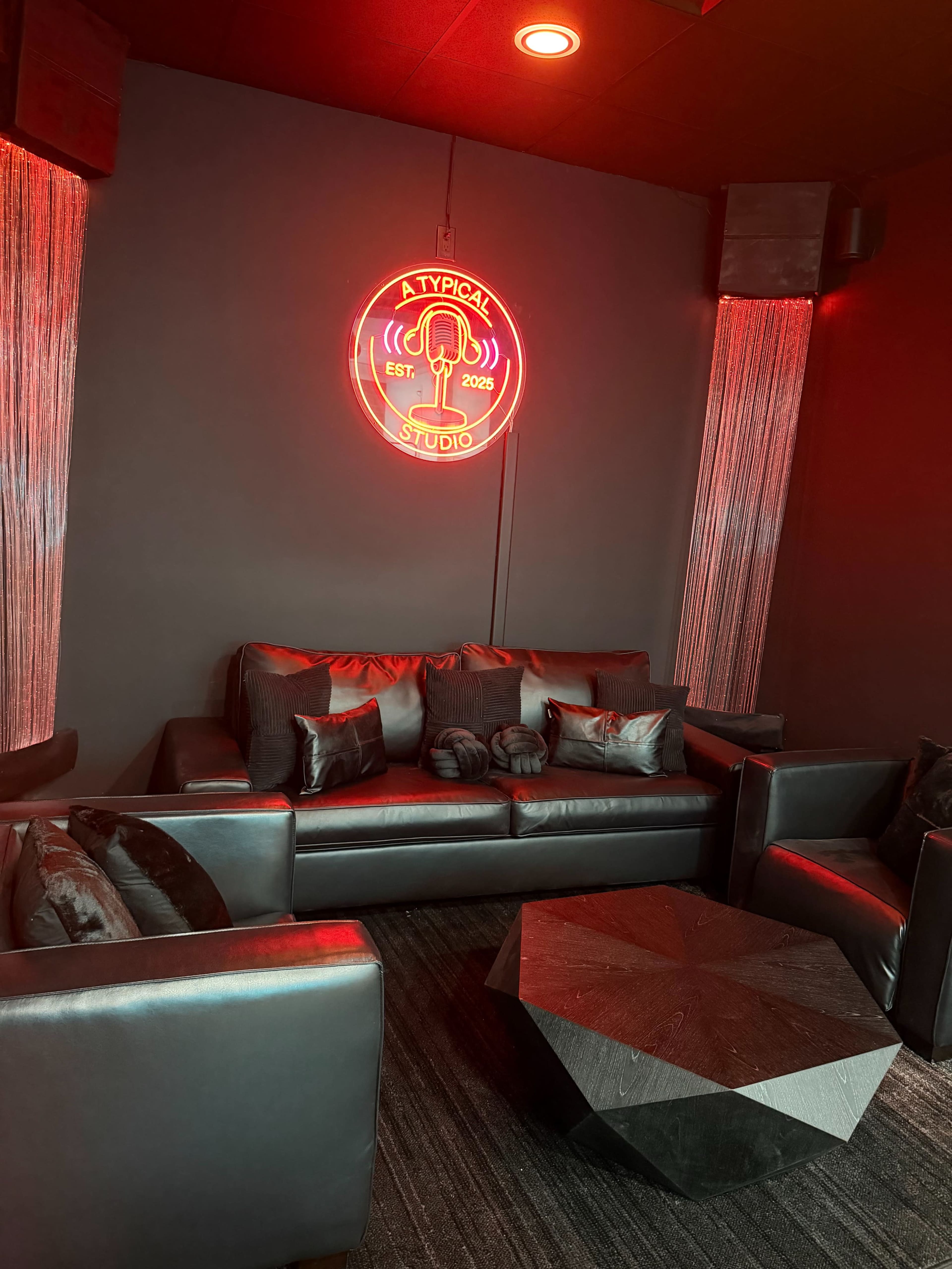 The image shows a modern lounge area with a black leather sofa, a geometric coffee table, and a neon wall sign that reads "A TYPICAL STUDIO EST. 2023."