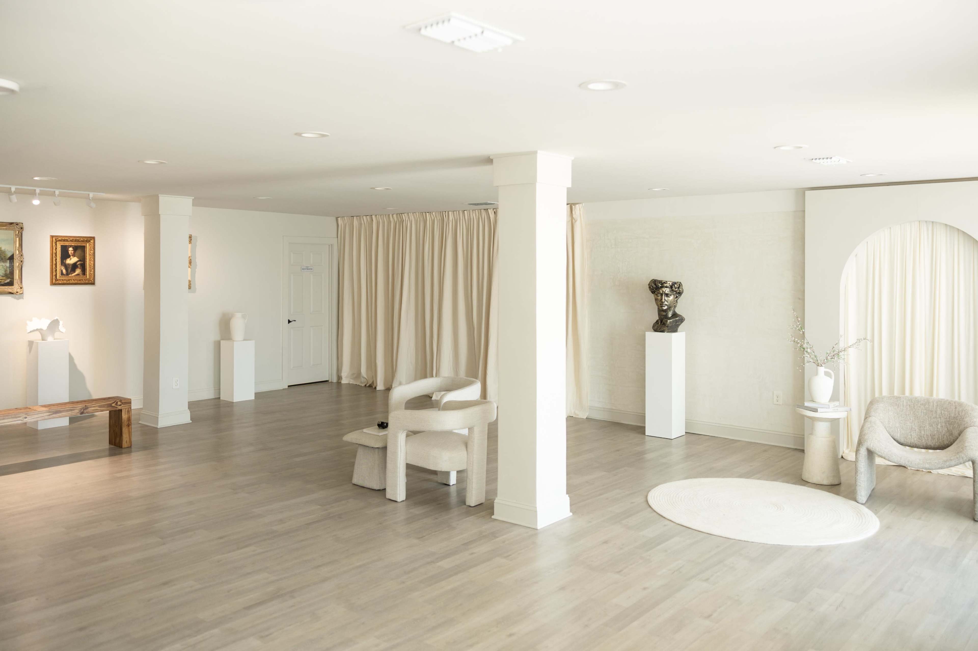 The image shows a spacious room with light-colored walls and floors, featuring minimalistic furniture, a couple of sculptures, and a large curtain on one side.