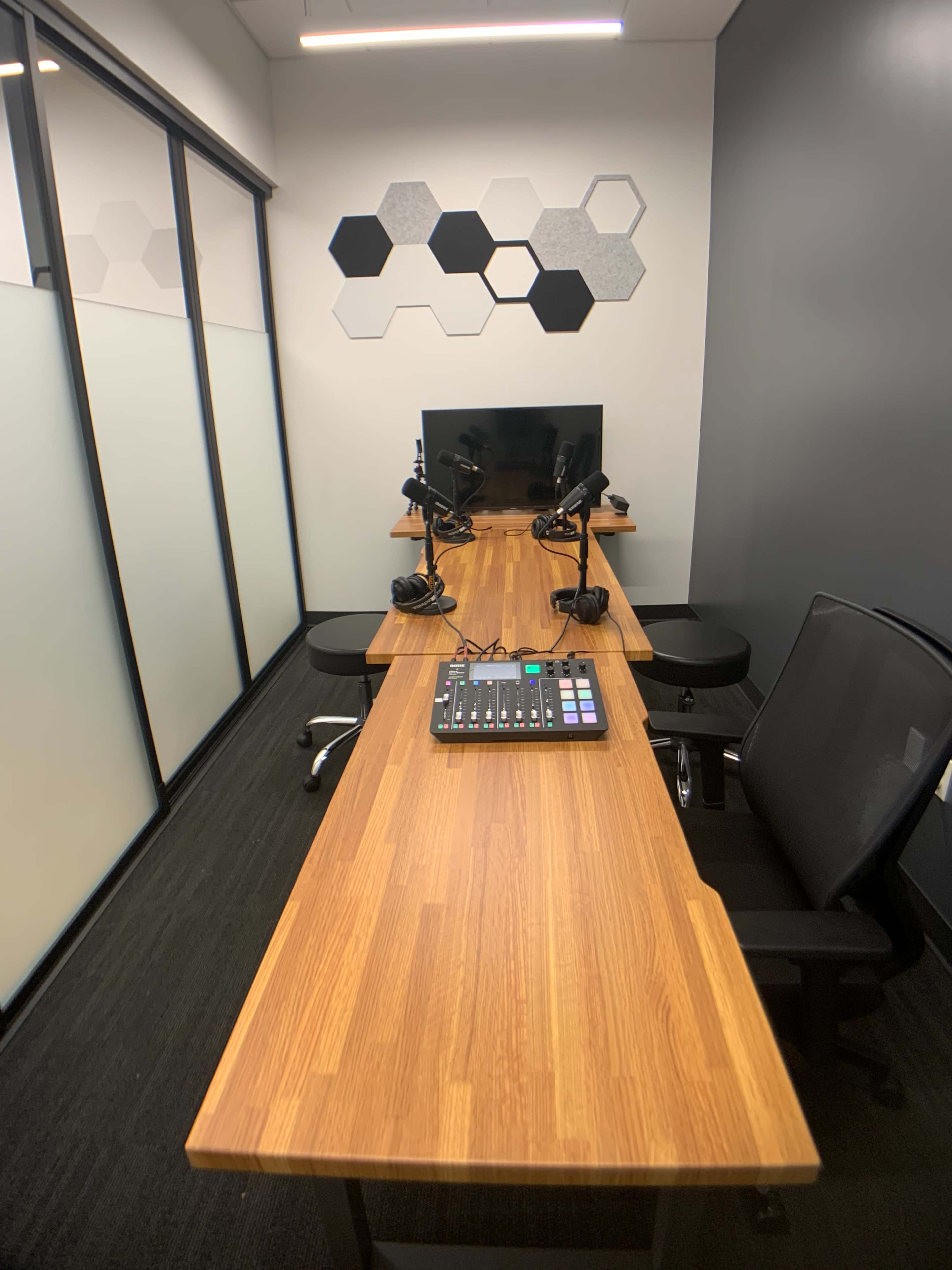A modern audio recording studio featuring a wooden table with two chairs, microphones, and a digital mixing console, with hexagonal wall decorations in the background.