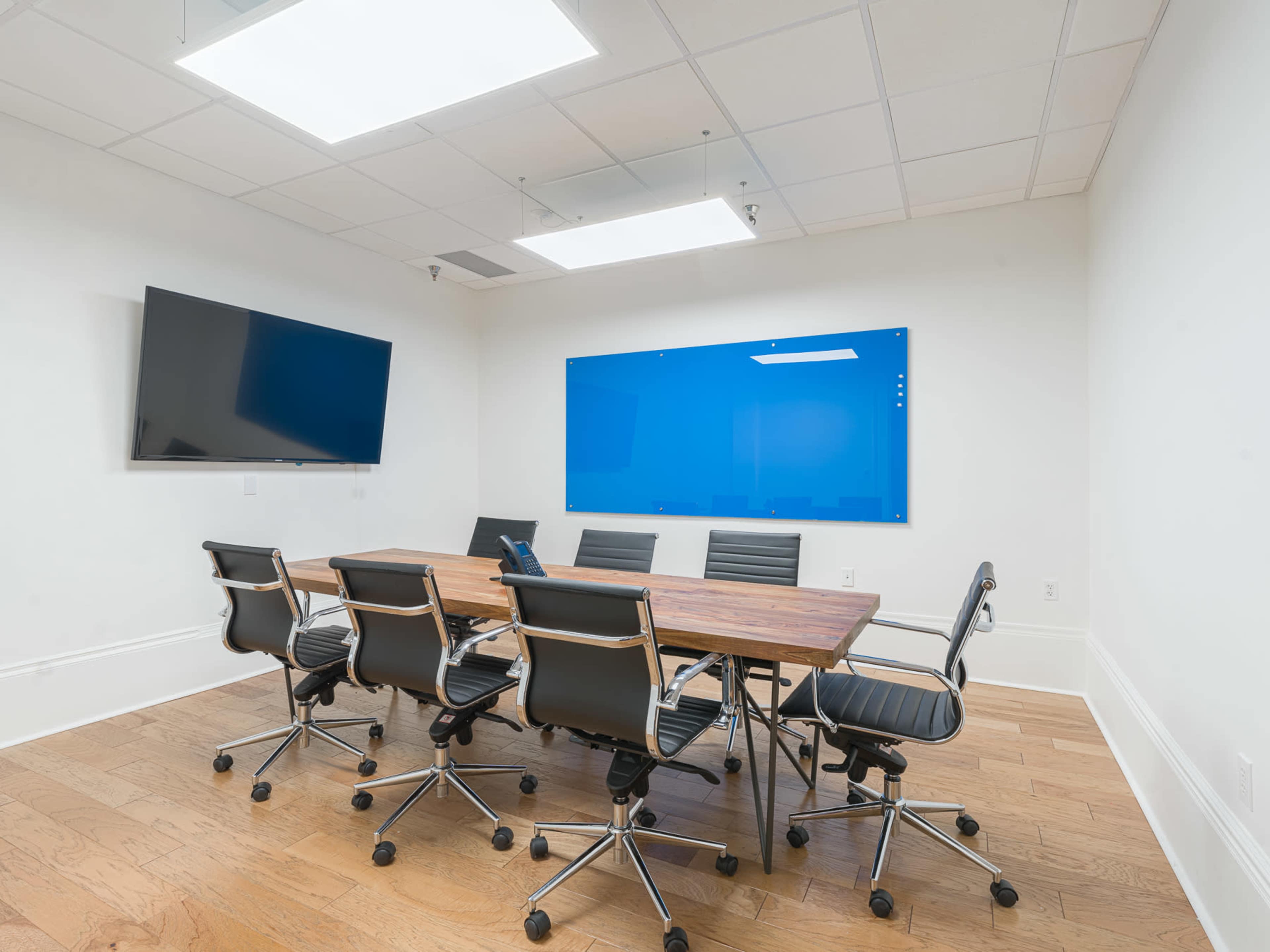 The image shows a modern conference room with a wooden table surrounded by black rolling chairs, two wall-mounted screens, and a blue whiteboard.