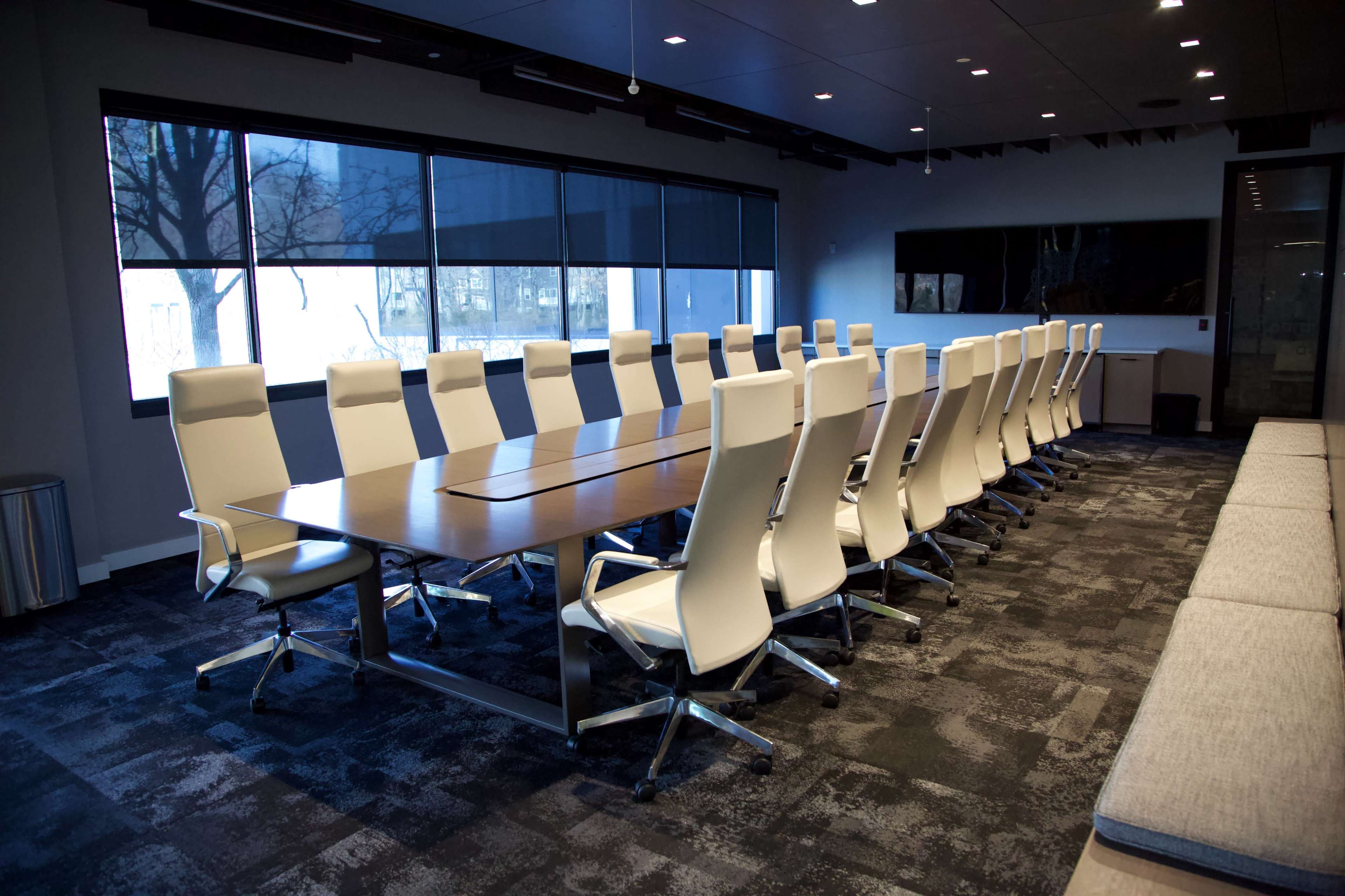 A large conference room features a long wooden table surrounded by white ergonomic chairs and large windows.
