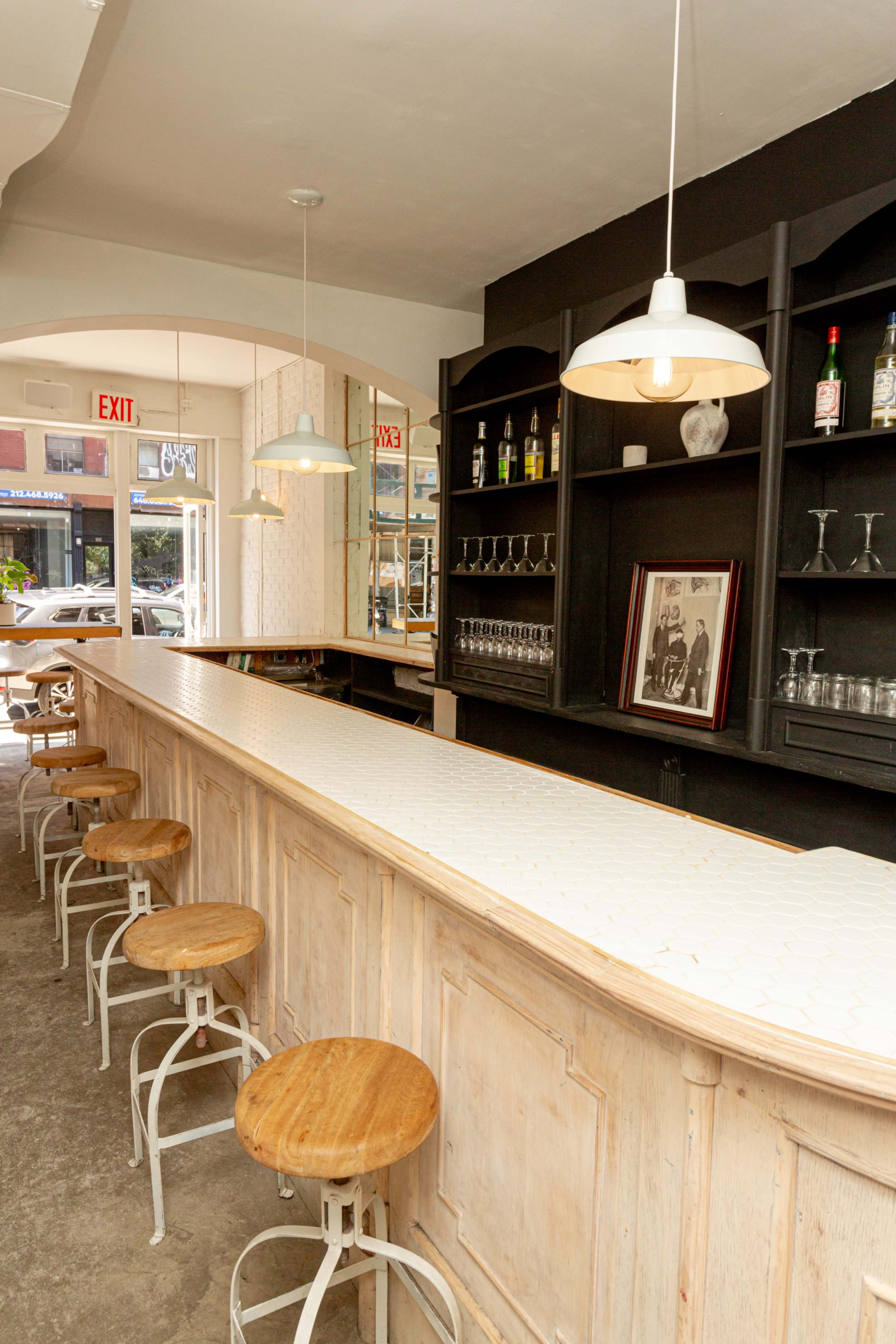 The image shows a light-colored bar with wooden stools arranged neatly in front of it, featuring mirrors and shelves displaying various bottles in a bright, inviting space.