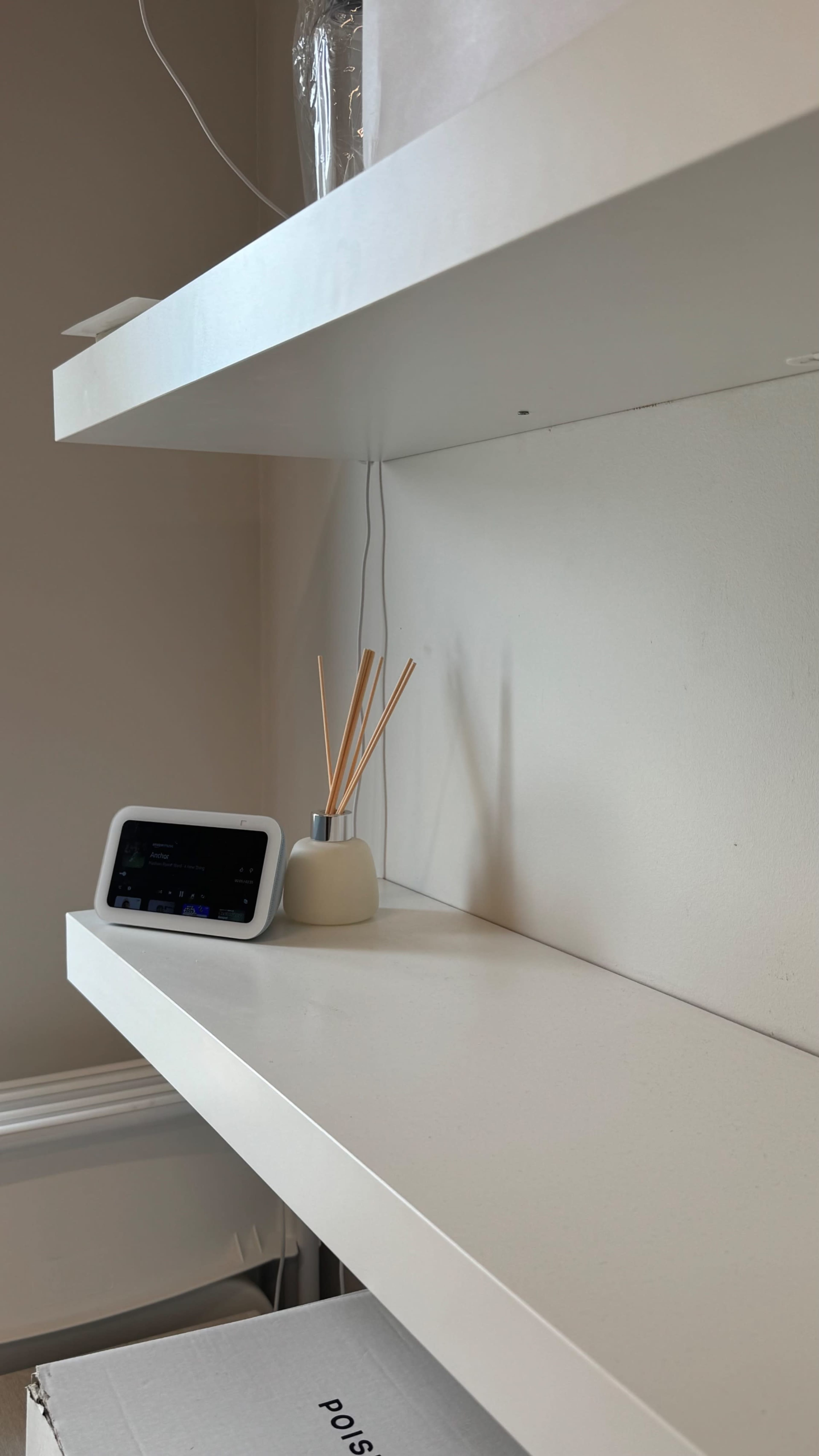 A minimalist shelf displays a smart speaker and a diffuser with wooden reeds against a plain wall.