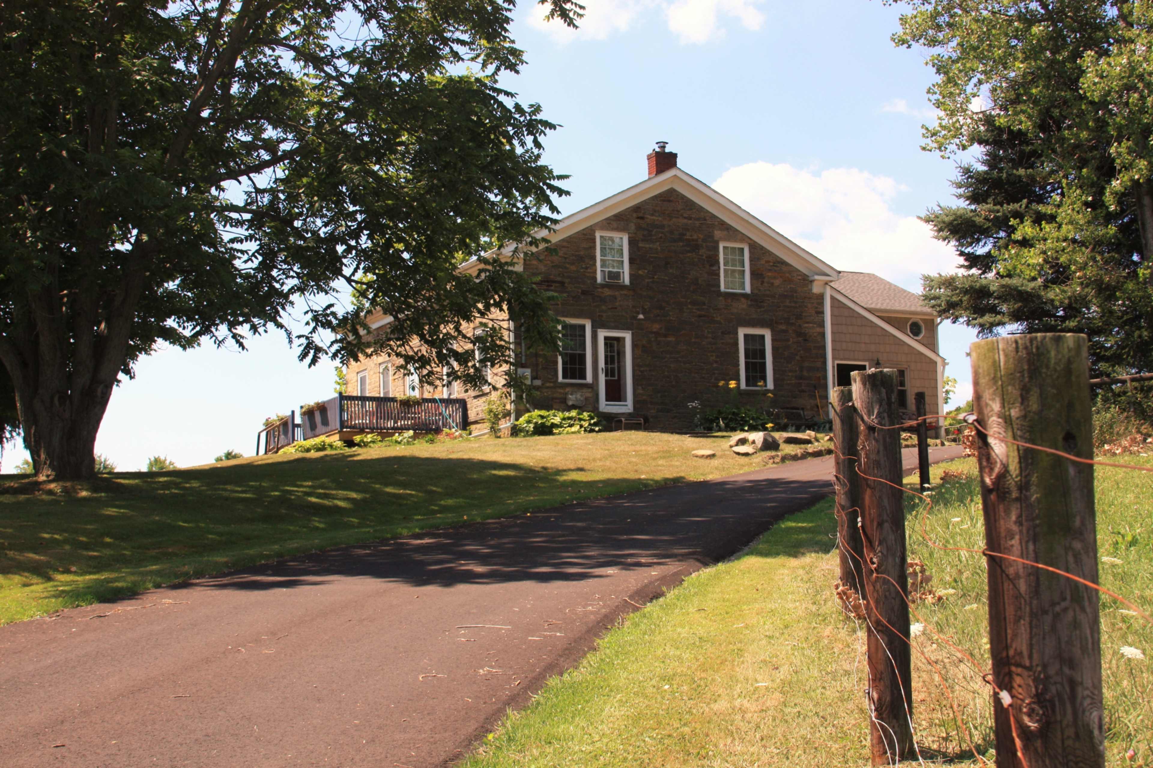 A stone house with a sloped driveway sits on a grassy hillside, surrounded by trees.