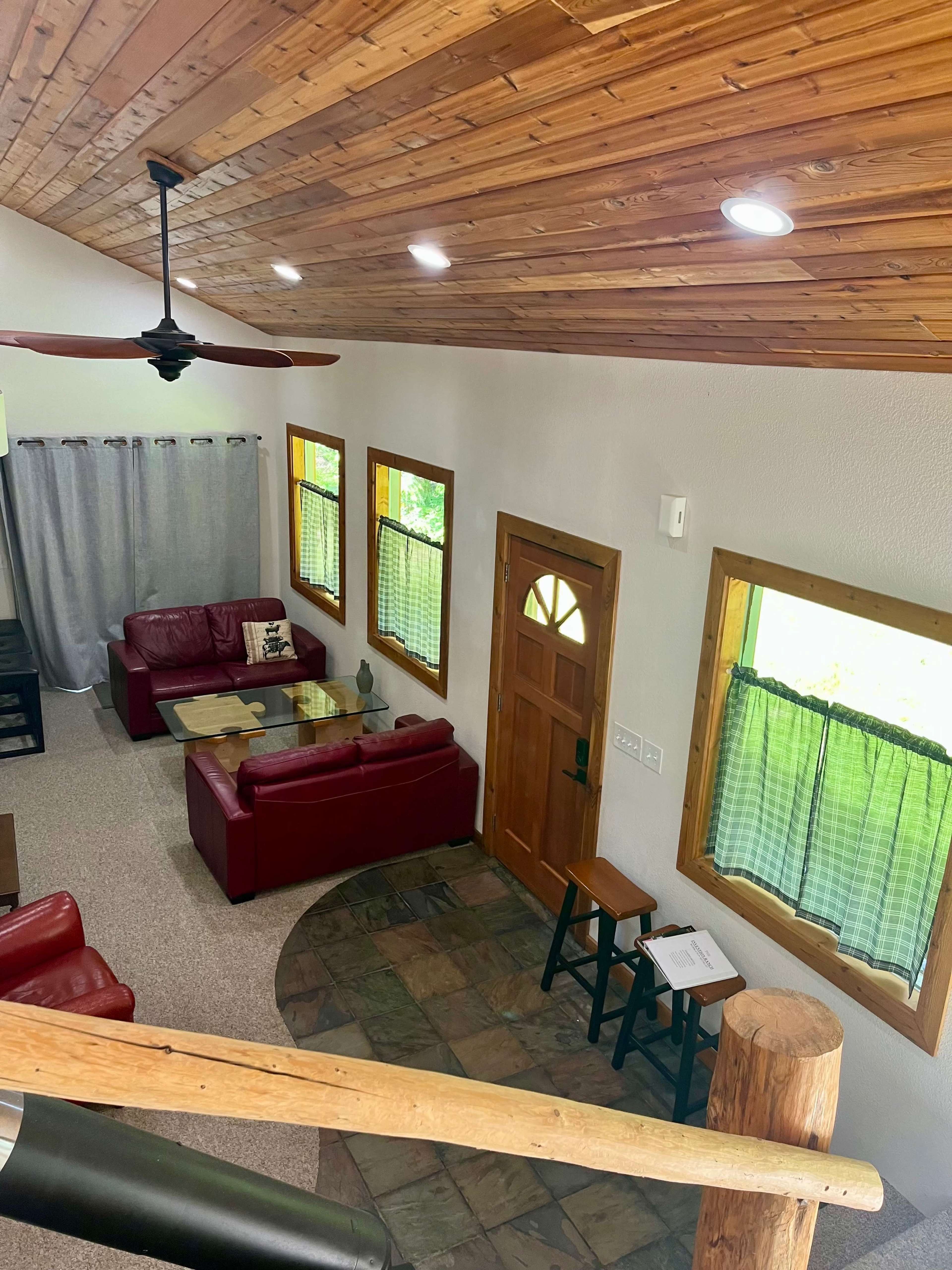 A cozy living area with red leather couches, a wooden coffee table, and large windows covered with curtains, all beneath a vaulted wooden ceiling.