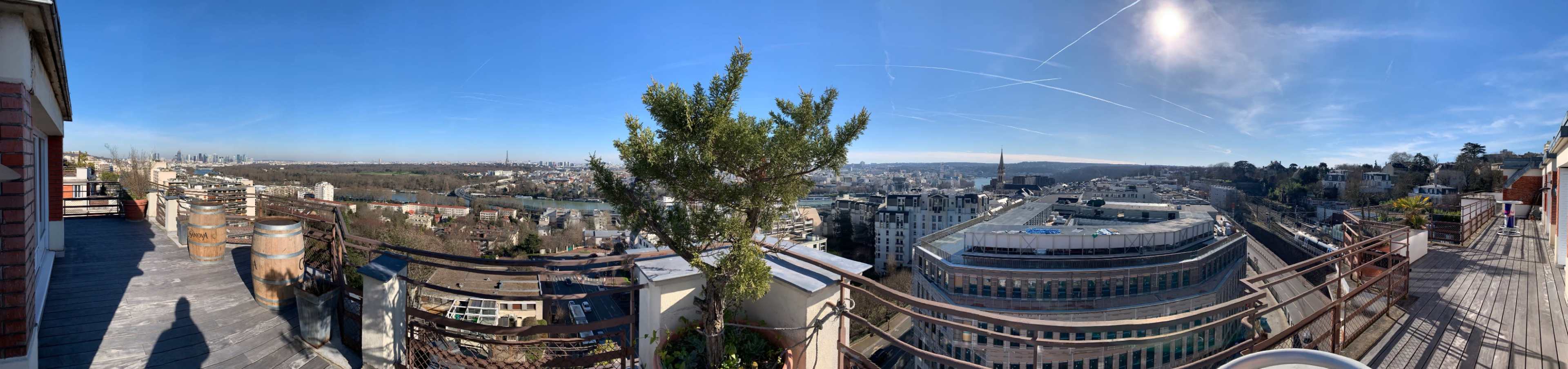 A wide panoramic view captures a rooftop scene overlooking a cityscape with buildings, trees, and a river under a clear blue sky.