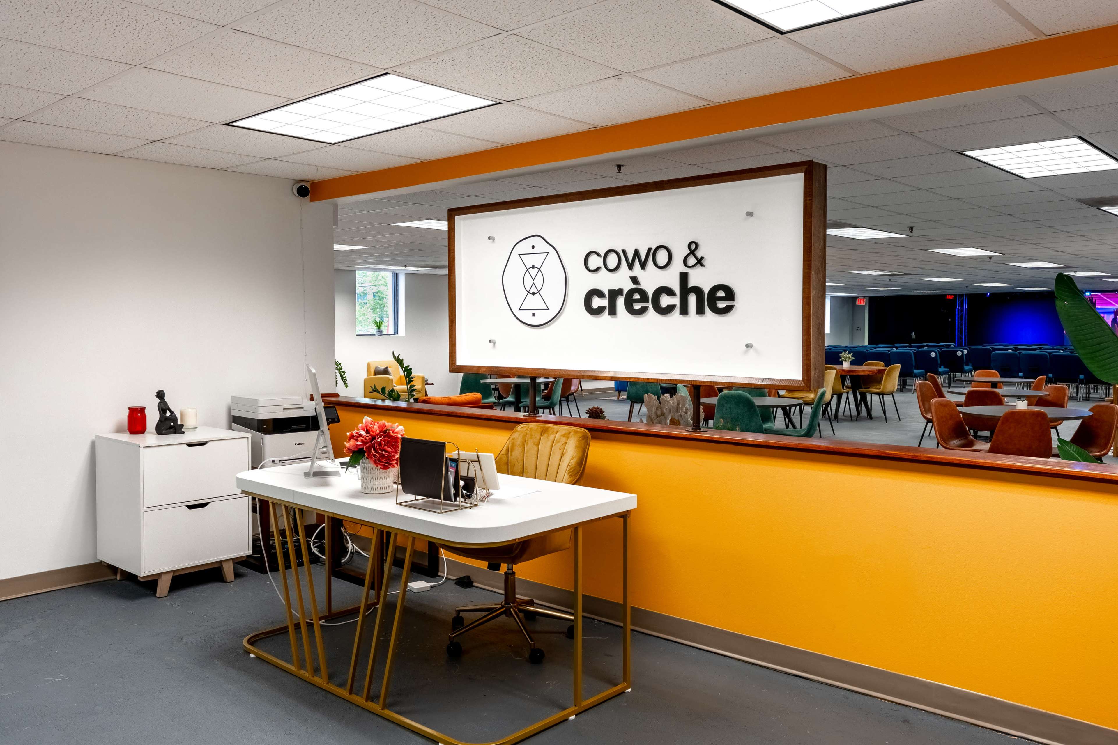 The image shows a bright coworking space featuring a reception desk with a sign reading "Cowo & crèche," along with colorful chairs and plants in the background.