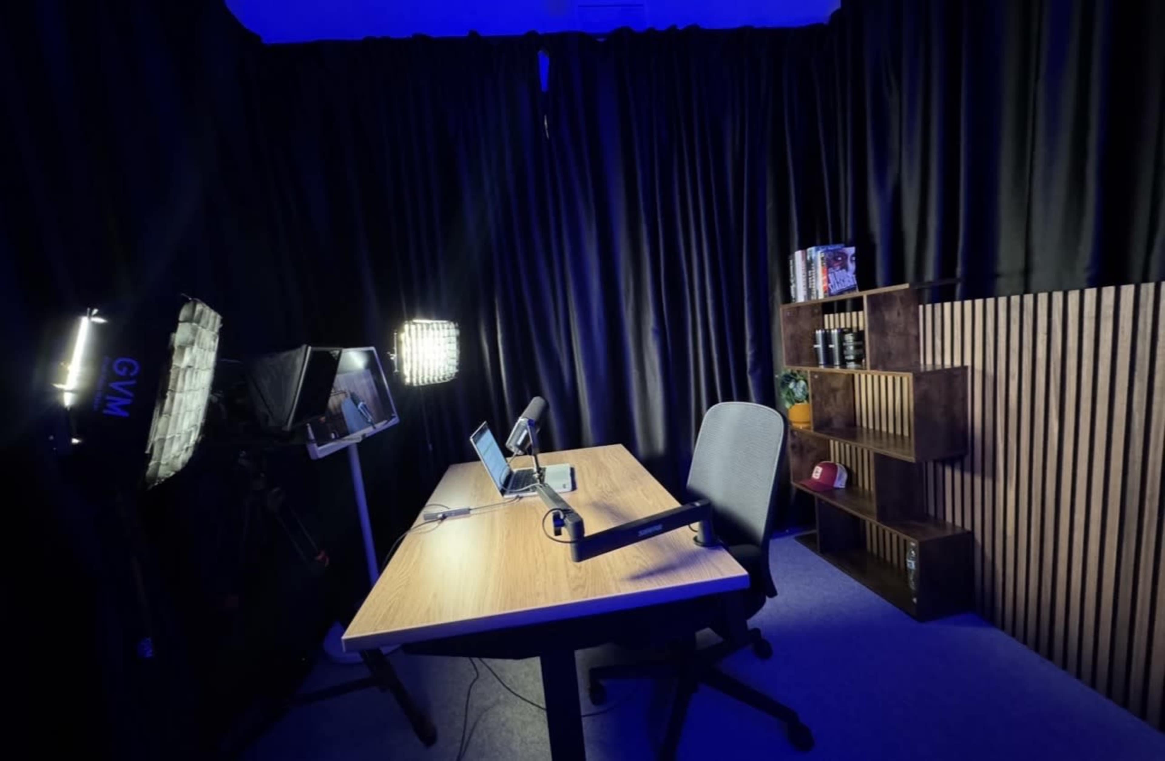 The image shows a dimly lit studio setup featuring a wooden desk, an office chair, a laptop, and a bookshelf against a backdrop of black curtains.