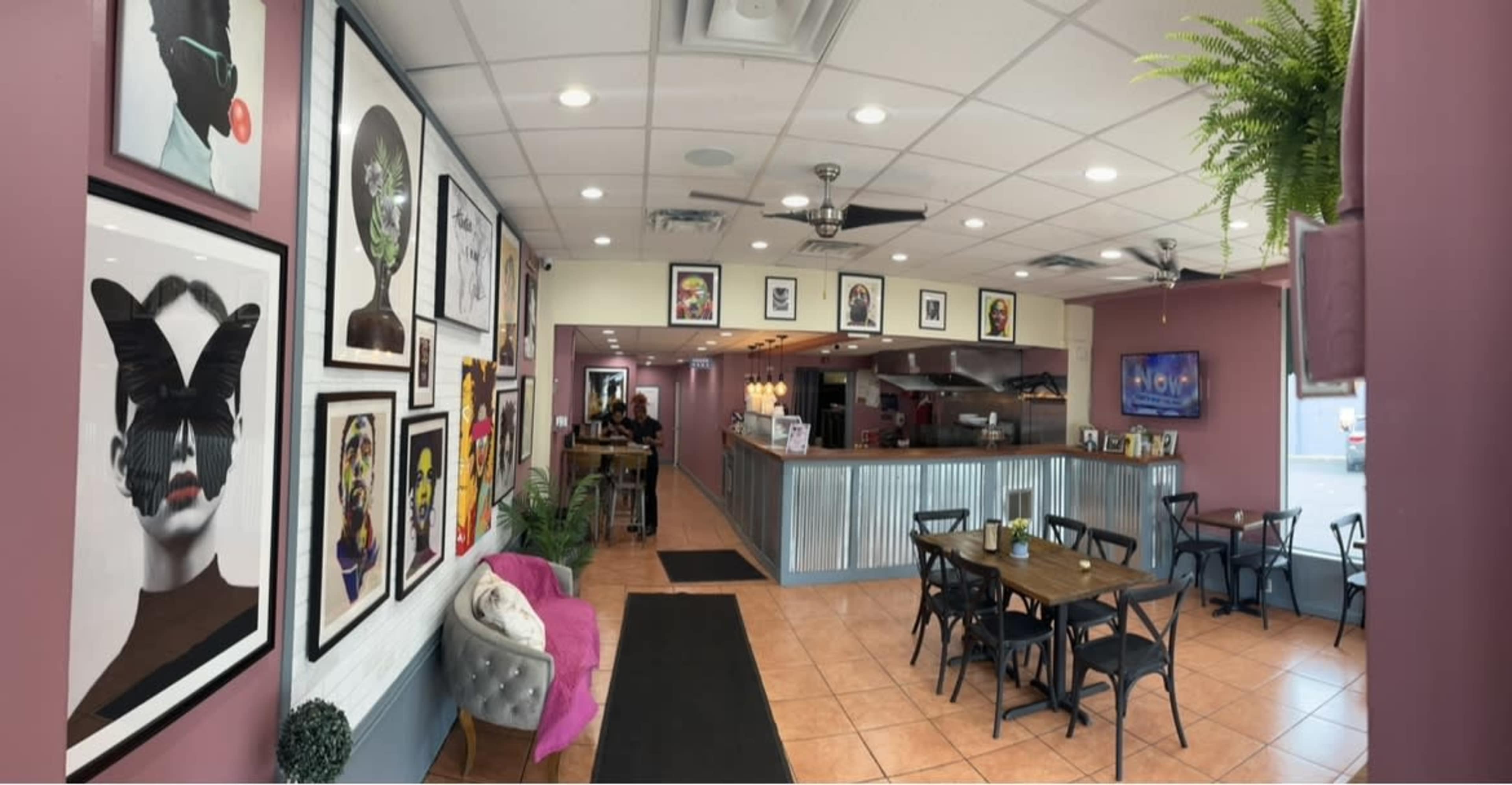 The interior of a cafe with a modern design, featuring colorful artwork on the walls, a seating area with tables and chairs, and a view of the kitchen at the back.