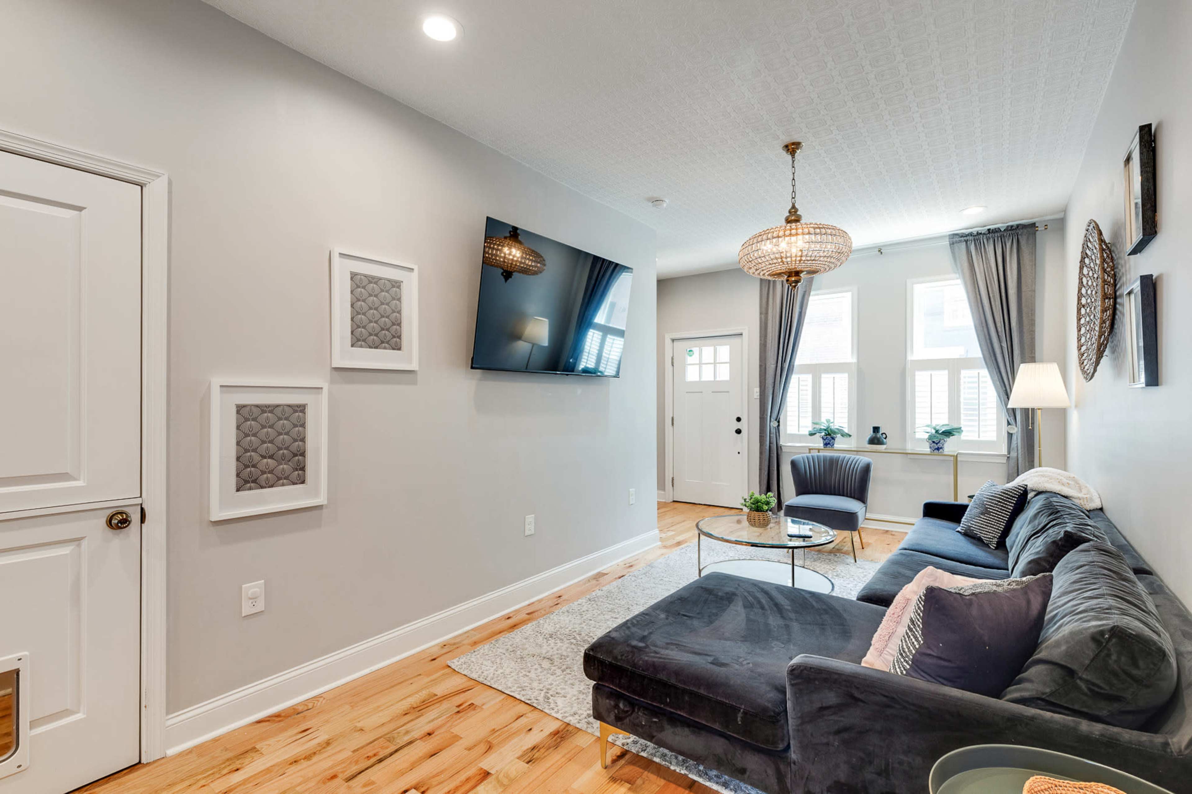 The living room features a dark sofa, a flat-screen TV mounted on the wall, and large windows with light drapes.