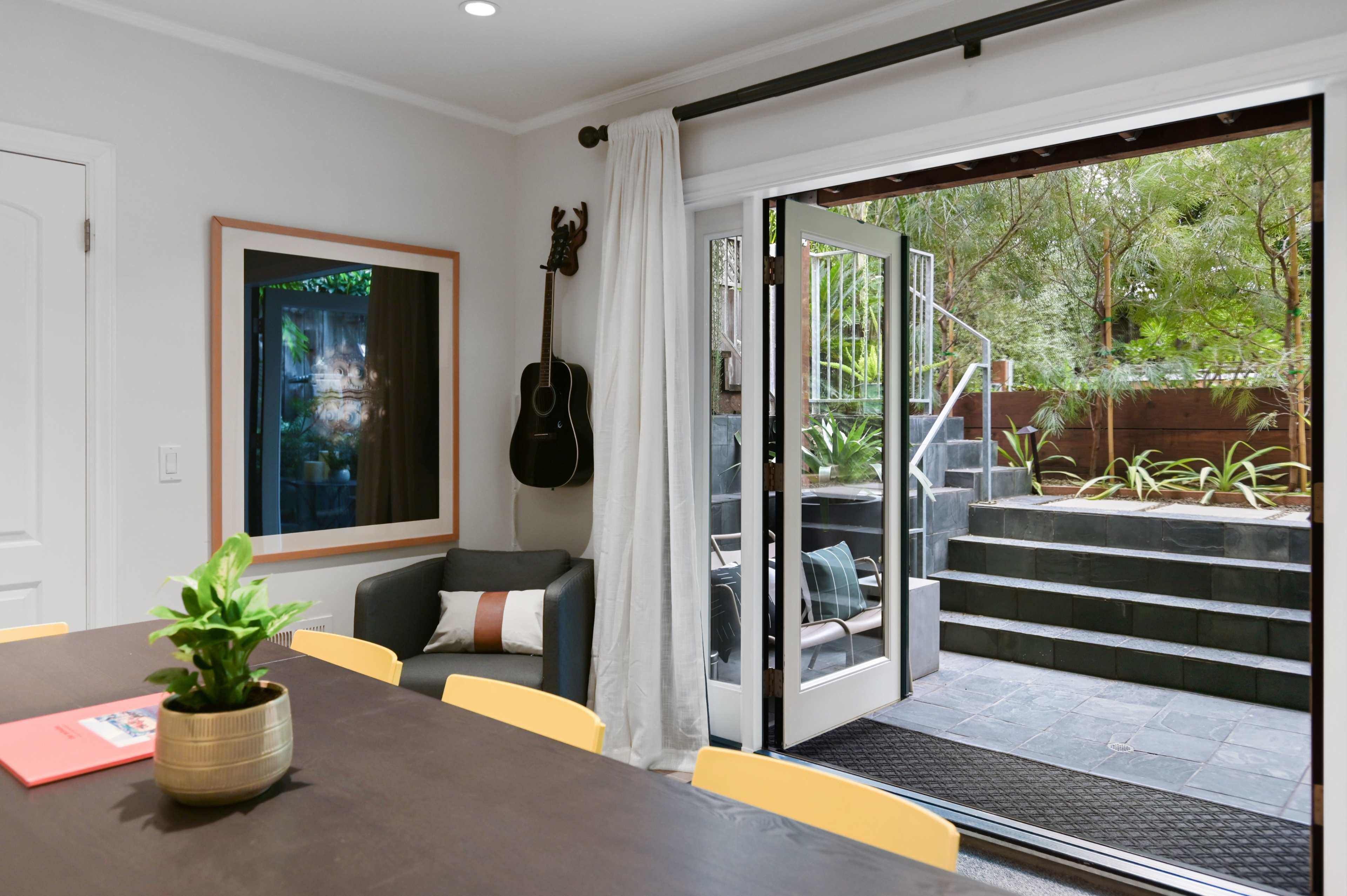 A modern dining area with a wooden table and yellow chairs adjacent to an open door that leads to a staircase and a garden.