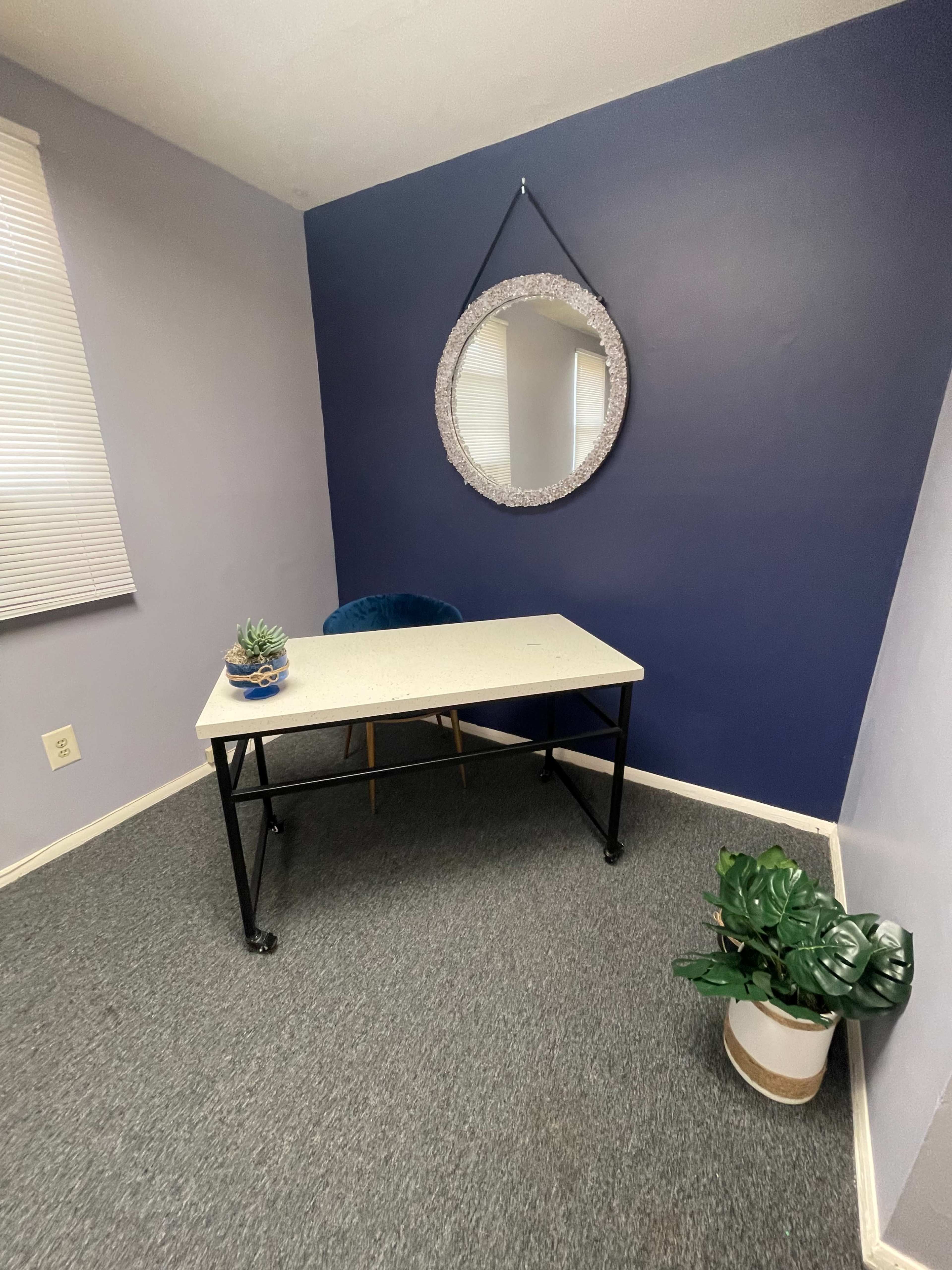 The image shows a small office space with a table, a chair, a decorative mirror on the wall, and a plant in a pot.