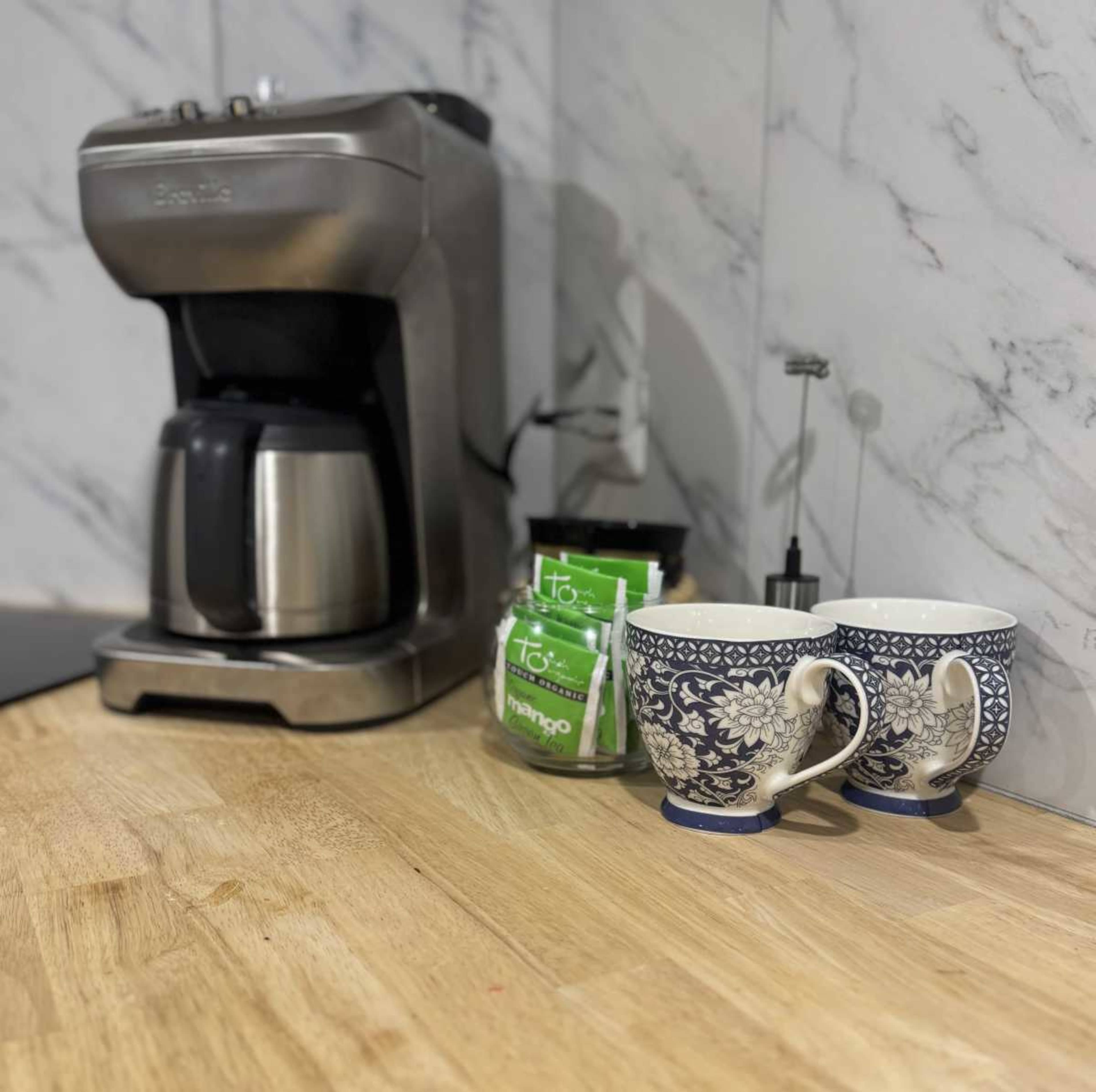 A coffee maker sits on a countertop next to two decorative mugs and several packets of tea.