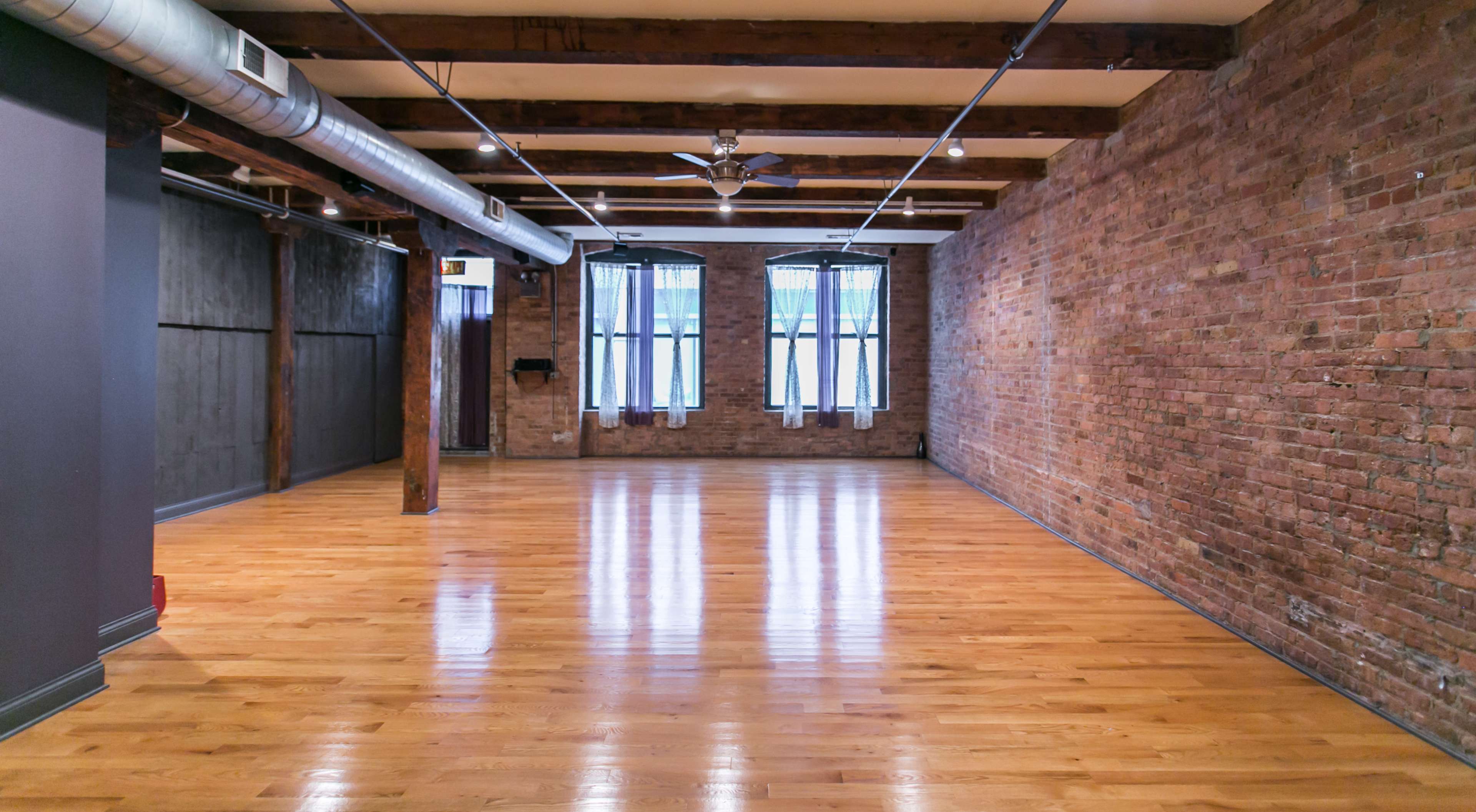 An empty room with exposed brick walls, wooden beams, and large windows allowing natural light to enter, highlighting the polished wooden floor.