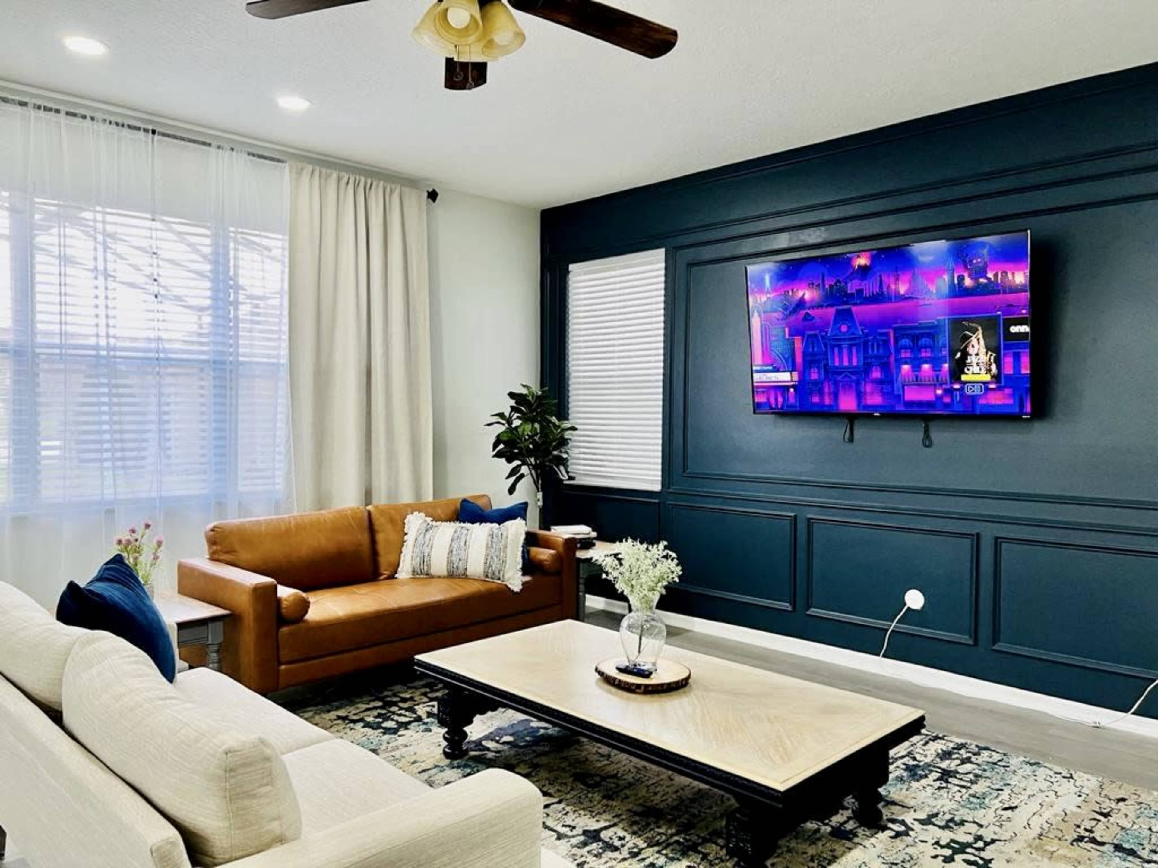 The living room features a light-colored sofa, a brown leather couch, a coffee table, and a television mounted on a dark blue accent wall.
