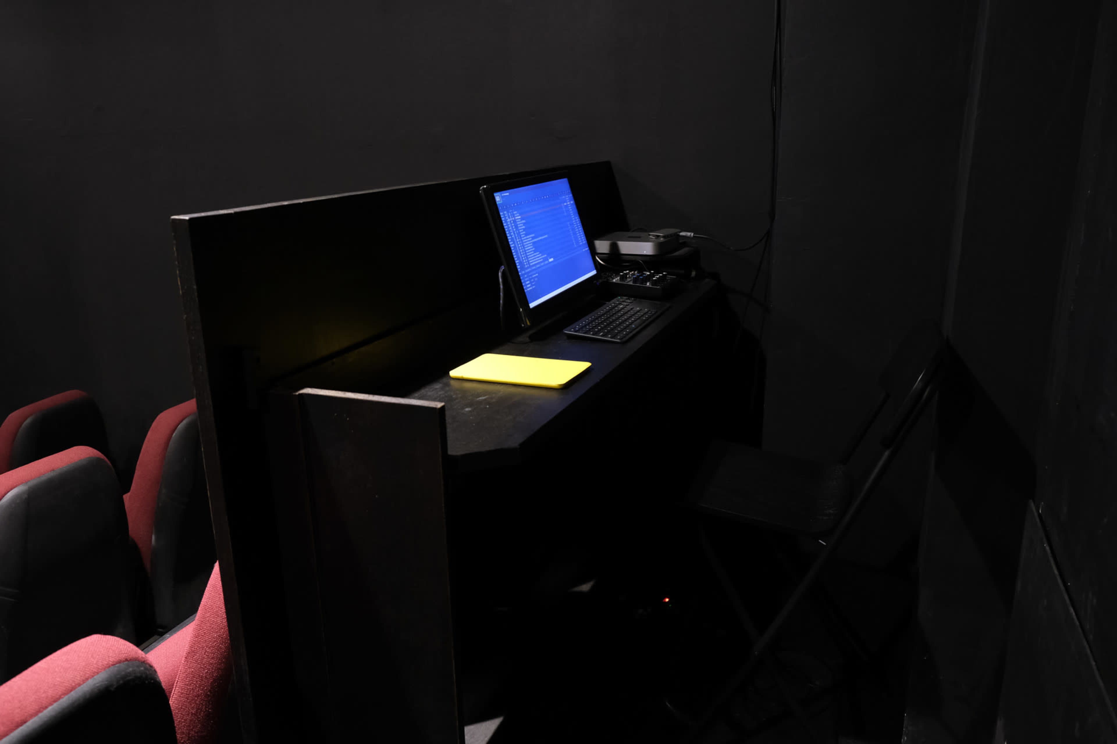 A dimly lit control booth features a desk with a computer, audio equipment, and a yellow notepad, adjacent to red theater seating.