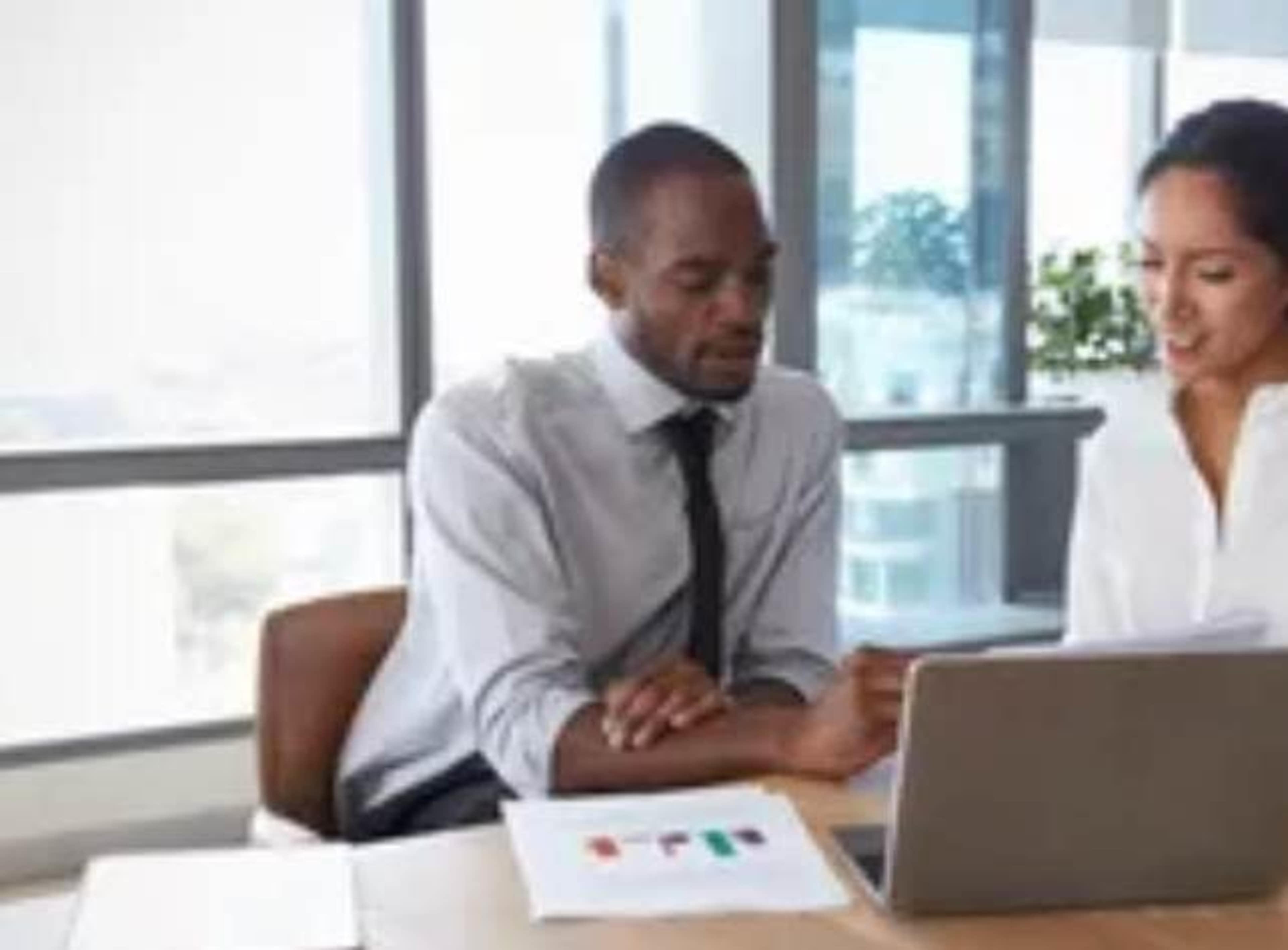 A man and a woman are discussing data on a laptop in a bright office with large windows.