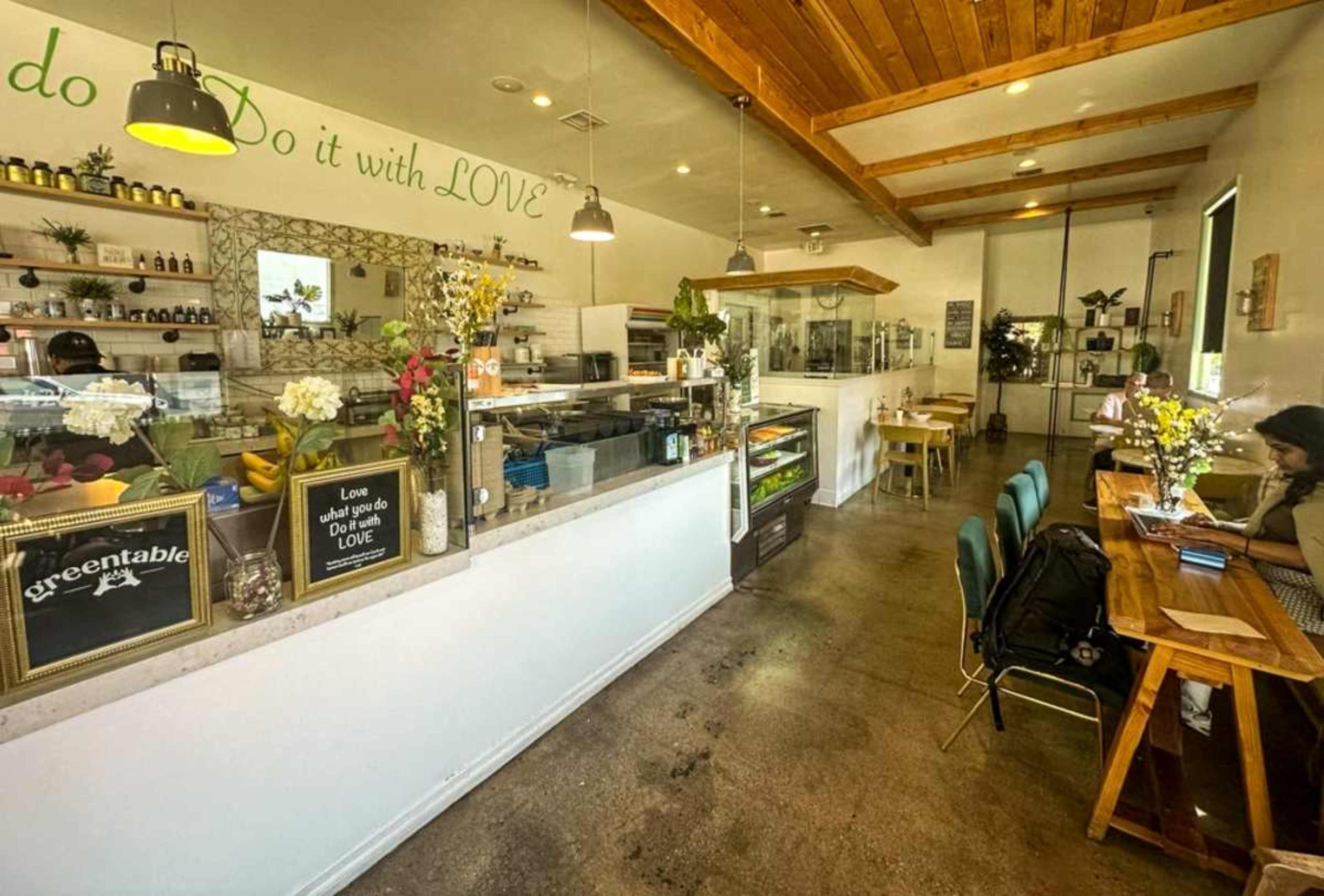 The image shows a bright café interior with a counter displaying various food items, tables for dining, and a person working on a laptop in the background.
