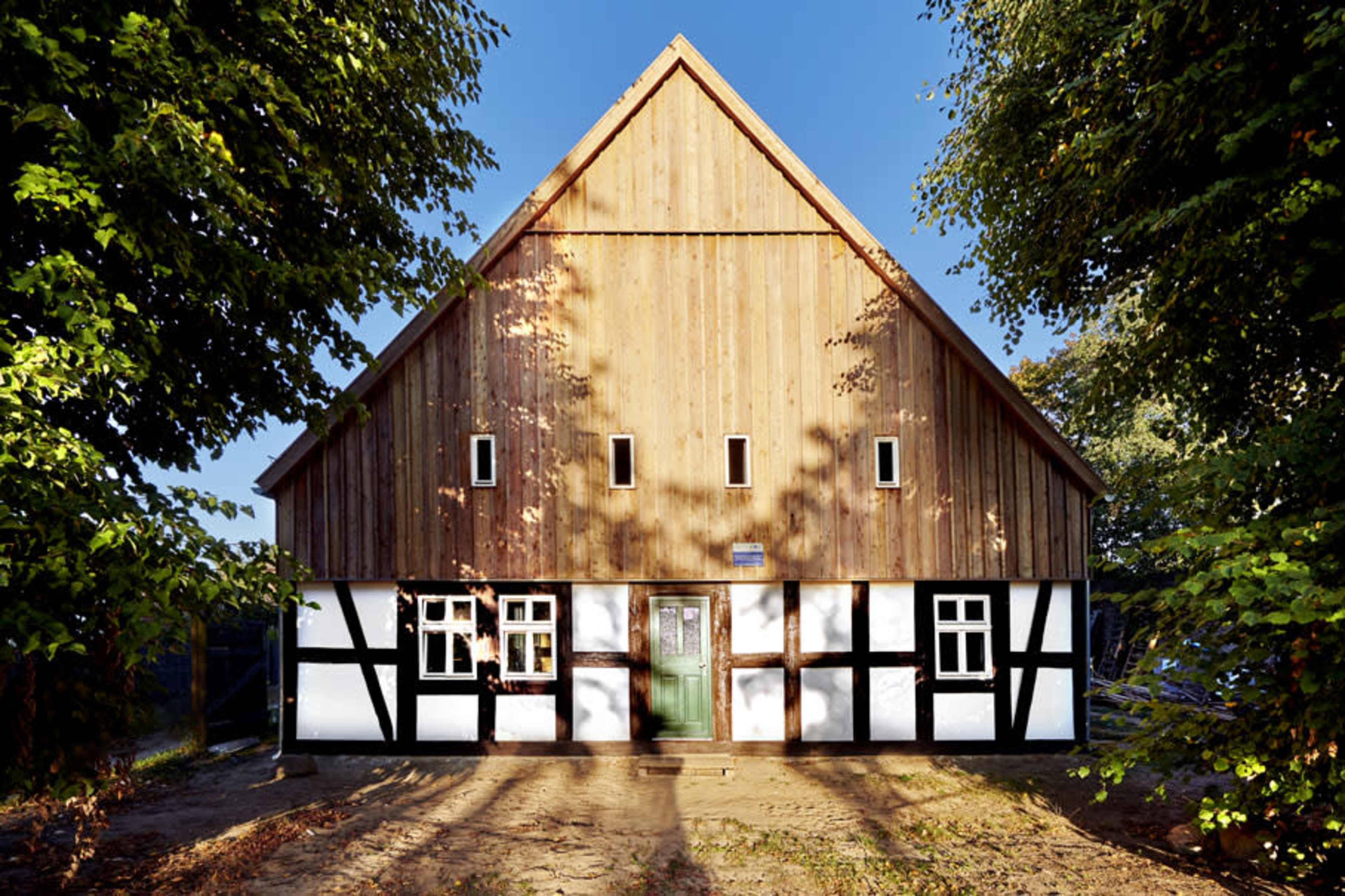 A traditional-style wooden house with a steeply pitched roof and a mix of wood and white plaster on the lower walls, surrounded by trees.