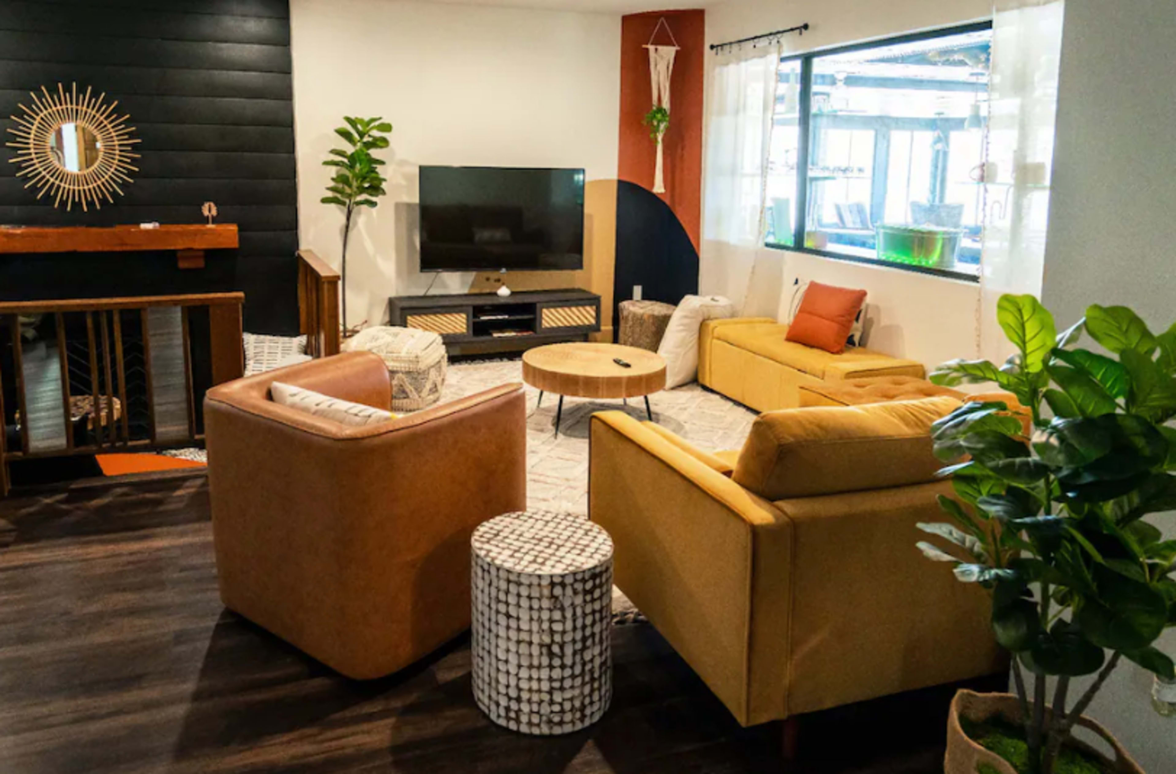 The image shows a modern living room with a television, two armchairs, a coffee table, a patterned rug, and decorative plants.
