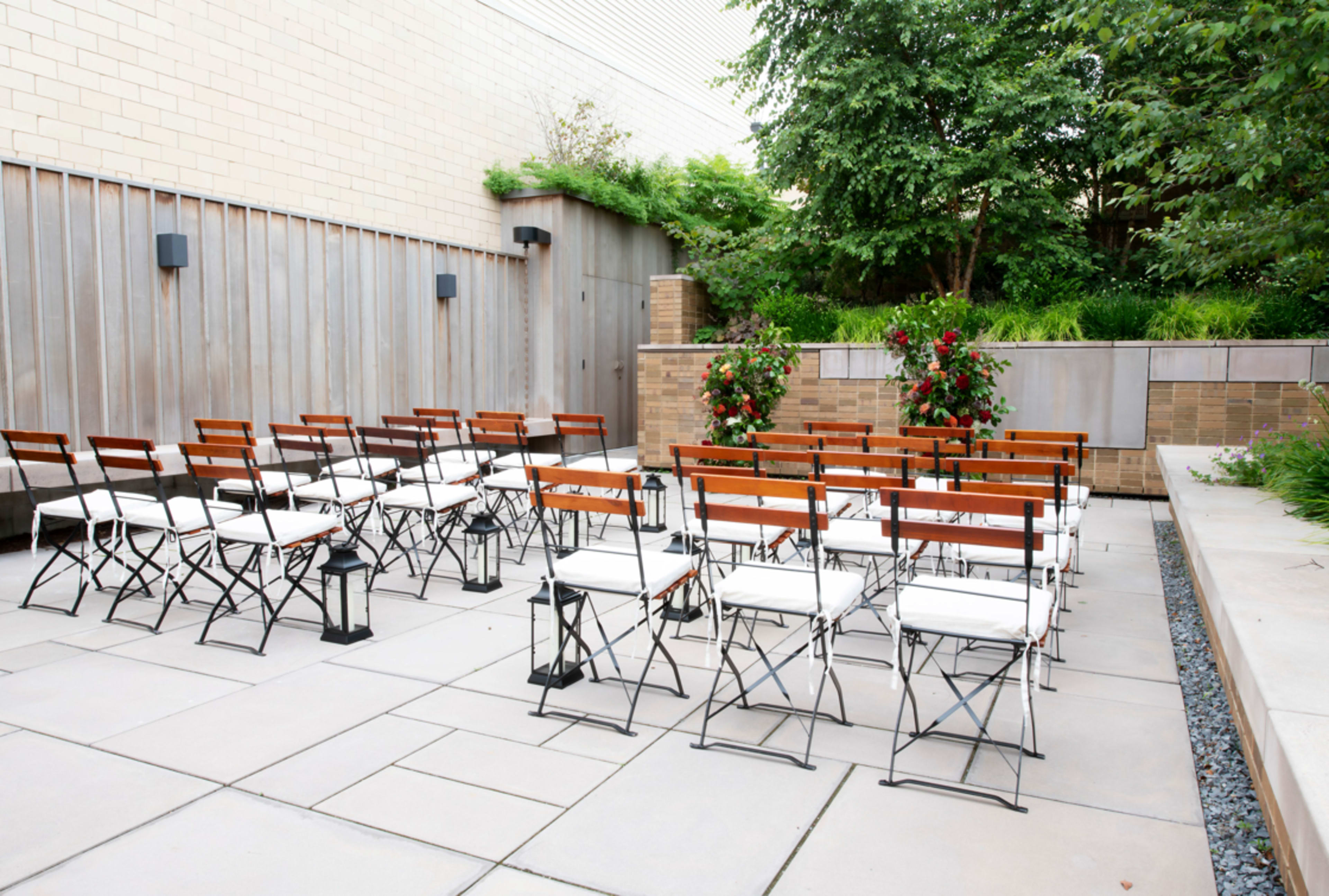 The image shows a set of wooden foldable chairs arranged in rows on a paved outdoor area surrounded by greenery.
