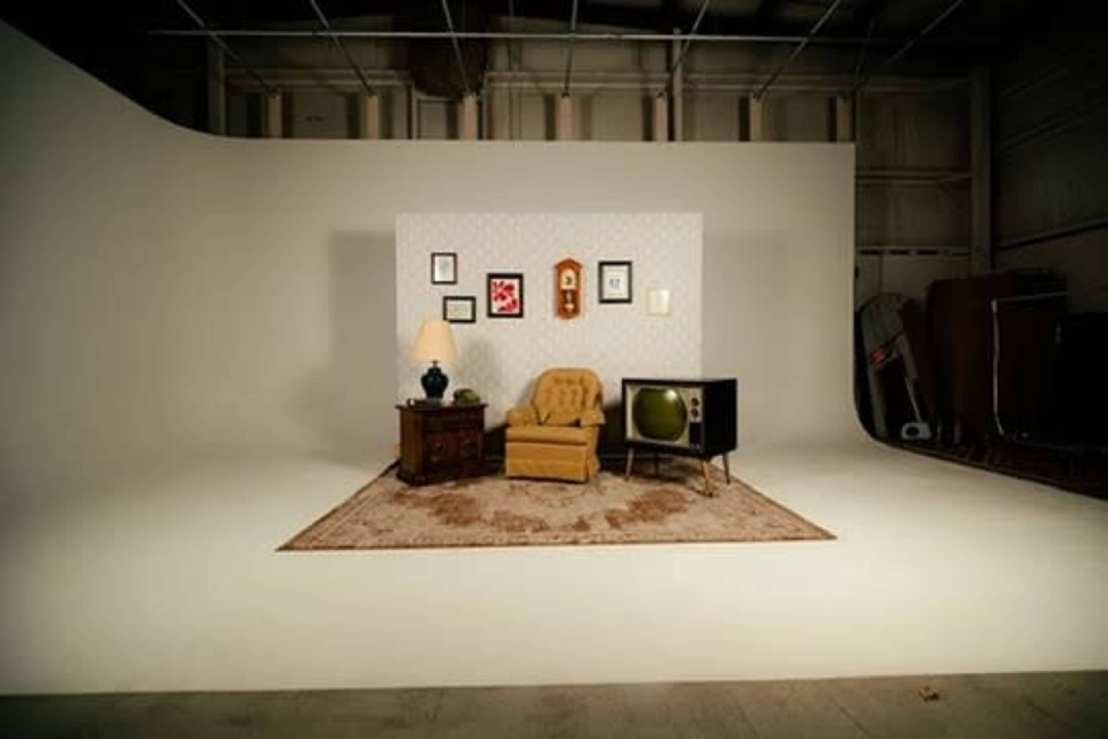 The image shows a sparse indoor scene featuring a yellow armchair, a wooden side table with a lamp, a vintage television, and a patterned wall with framed pictures, all set against a plain backdrop.