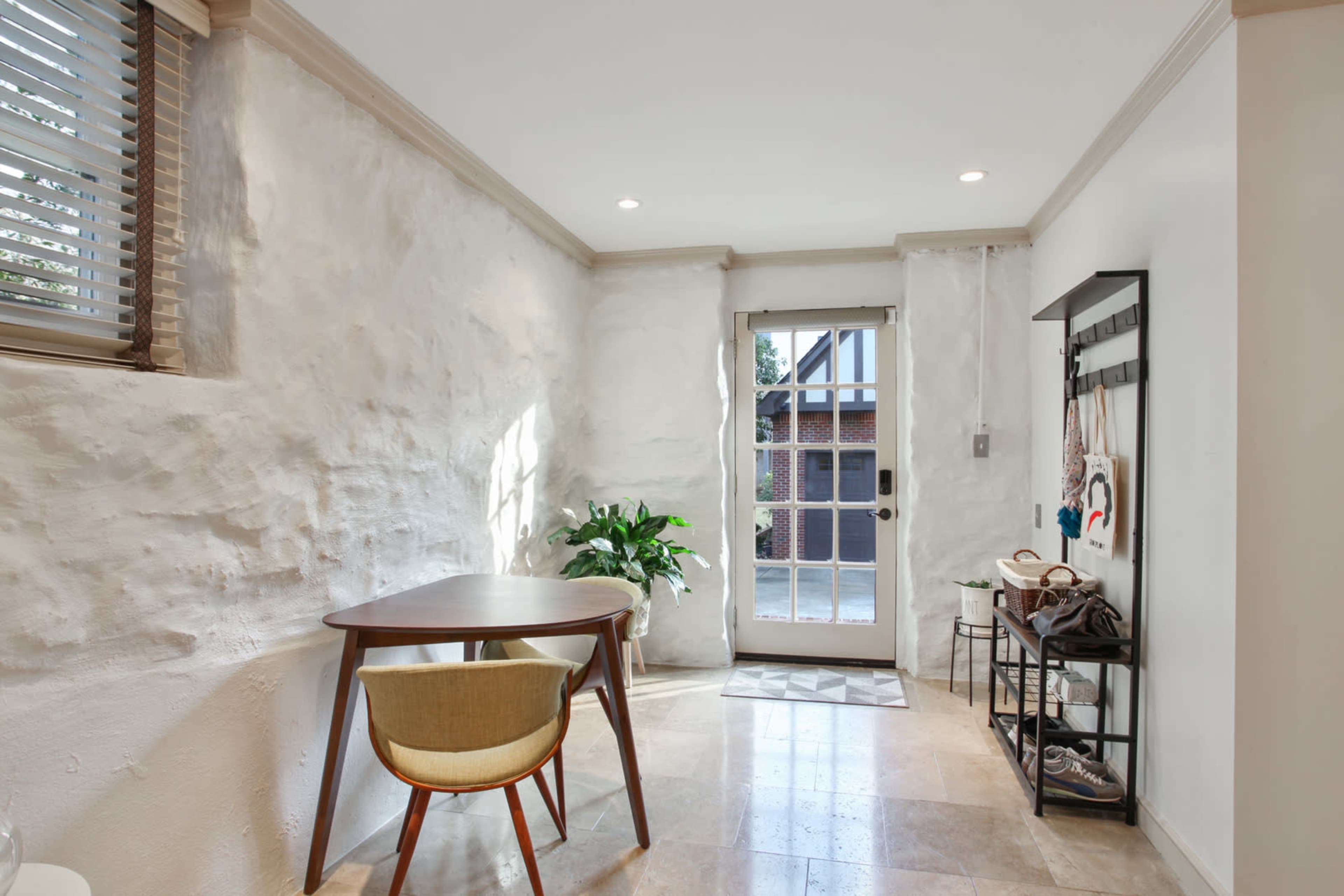The image shows a small, brightly lit room with a table and chair, a potted plant, and a door leading outside.