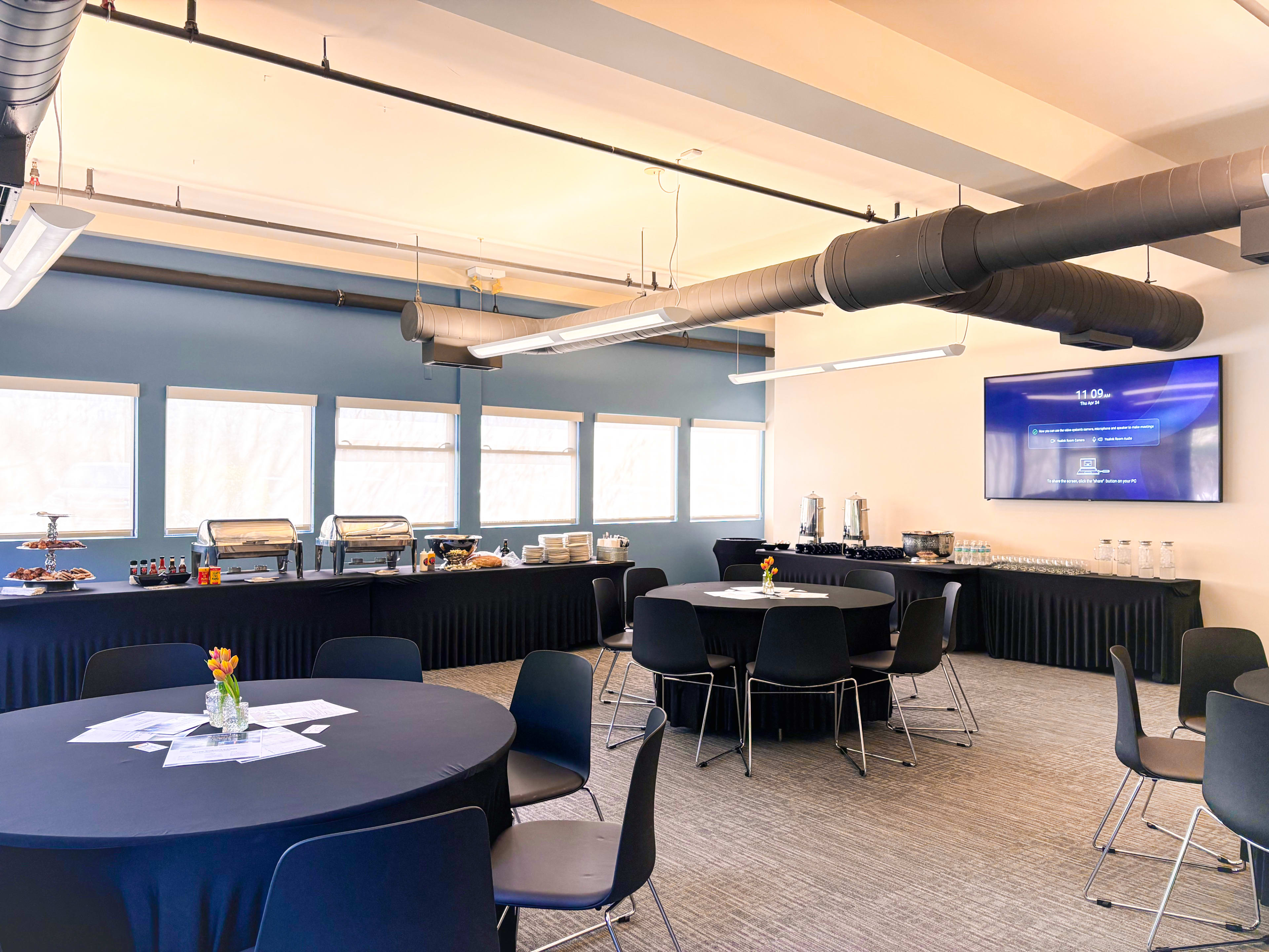 A spacious meeting room features round tables with black tablecloths, a buffet setup, and large windows allowing natural light to enter.