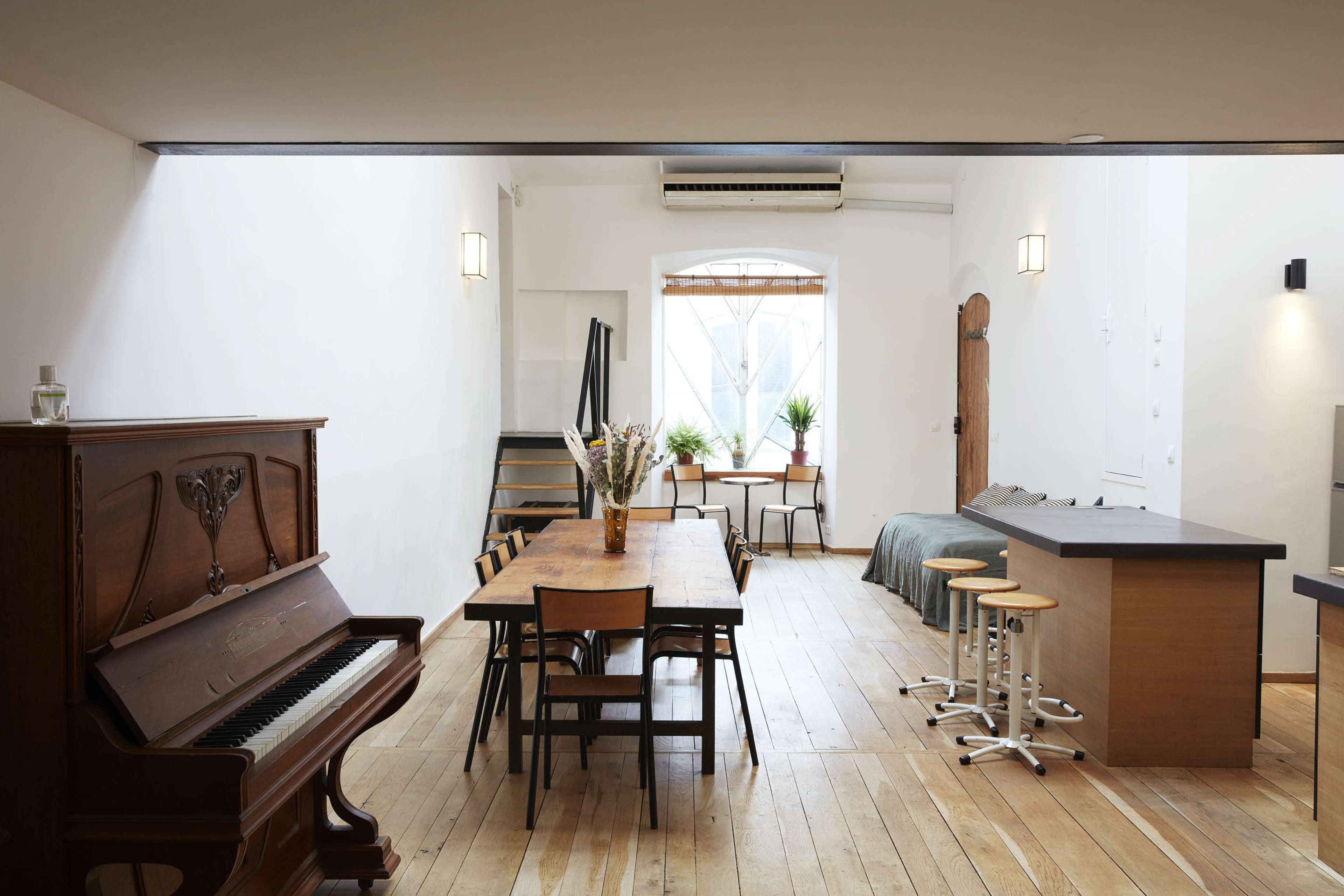 The image shows a spacious, modern interior with a wooden dining table, a piano, and a kitchen area featuring bar stools, all illuminated by natural light from a window.