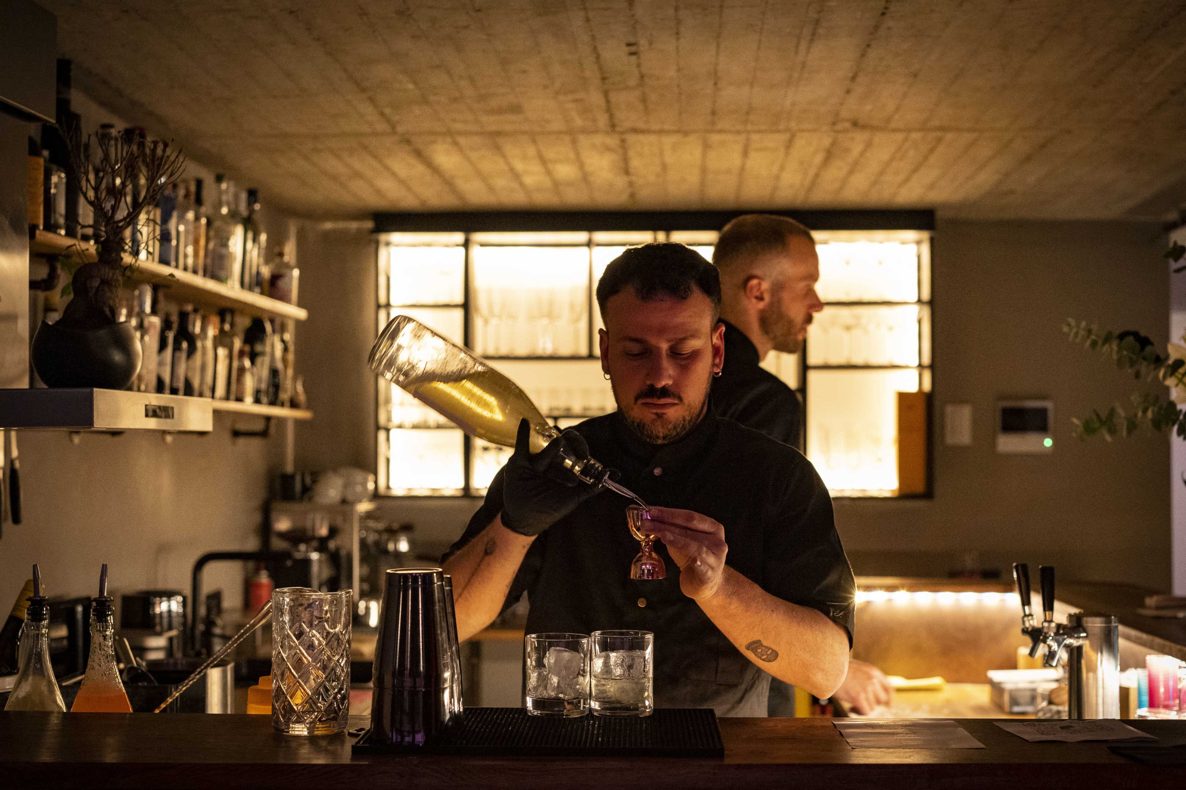A bartender in a black shirt pours a drink into two glasses at a dimly lit bar while another bartender works in the background.