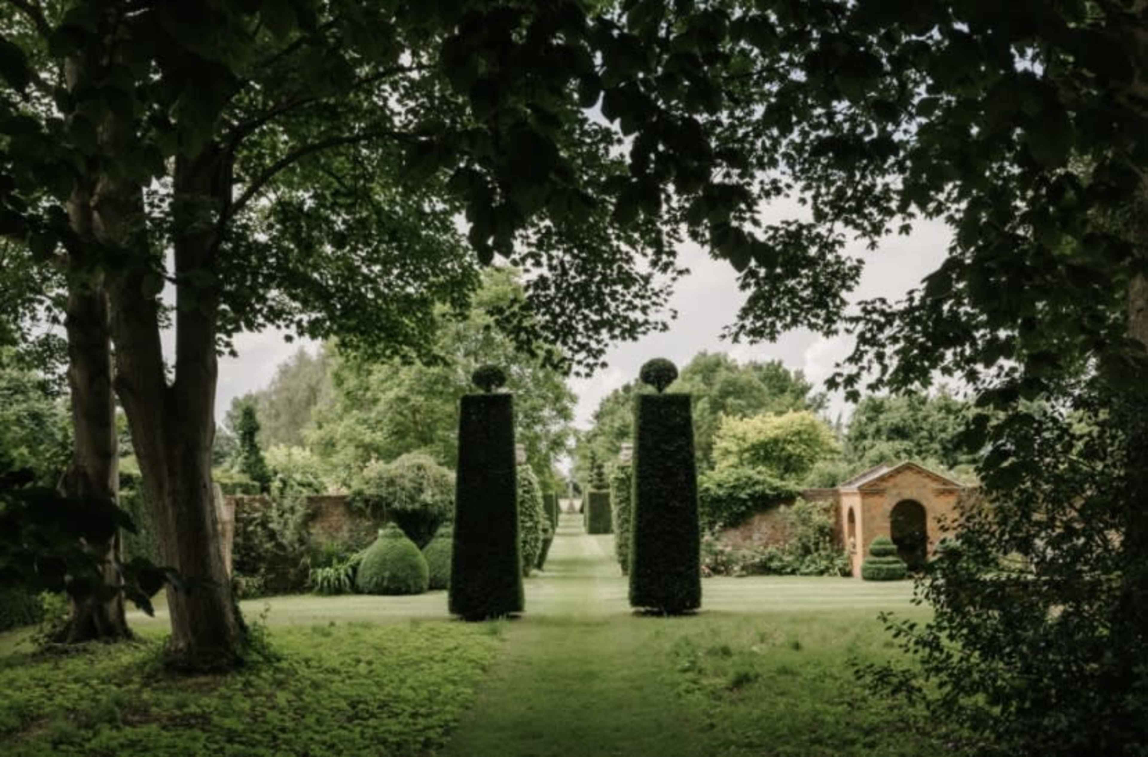 The image depicts a landscaped garden featuring tall, neatly trimmed hedges and symmetrical trees flanking a pathway that leads to a brick structure in the background.