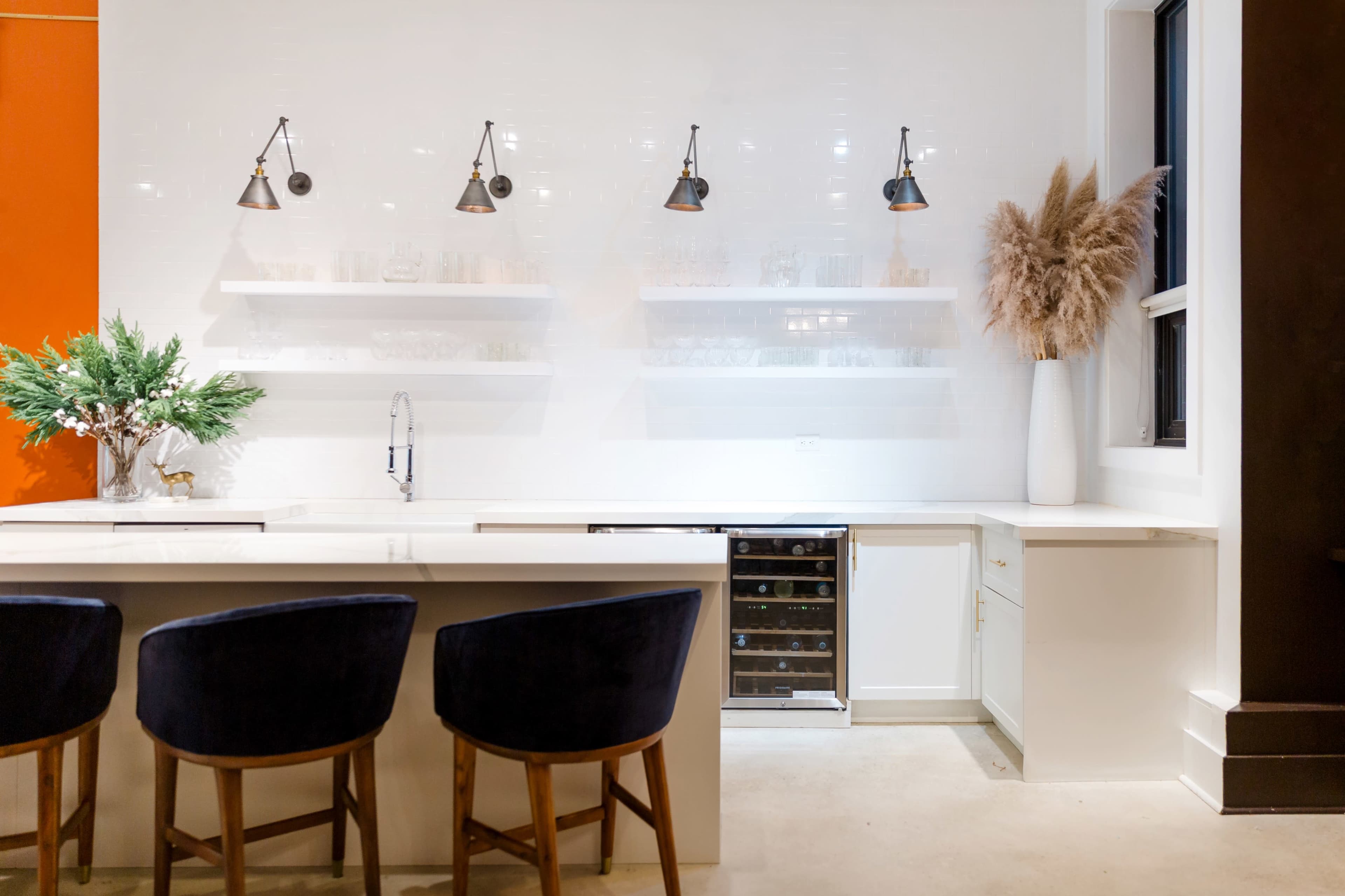 The image shows a modern kitchen with a white countertop, dark wood bar stools, sleek shelving, and decorative plants in vases.