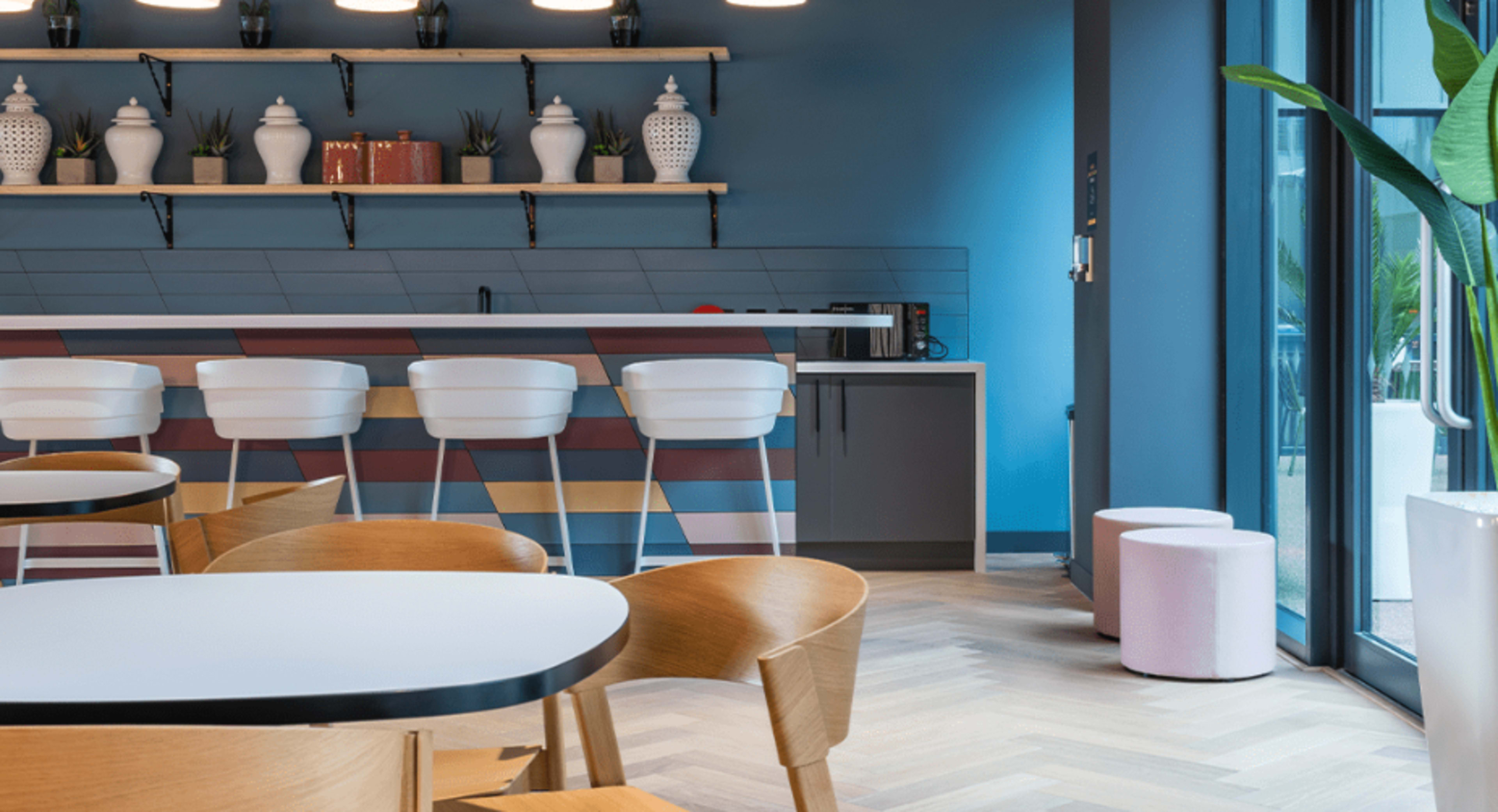 A modern kitchen area with a colorful bar counter, white stools, wooden tables, and a plant near a large window.