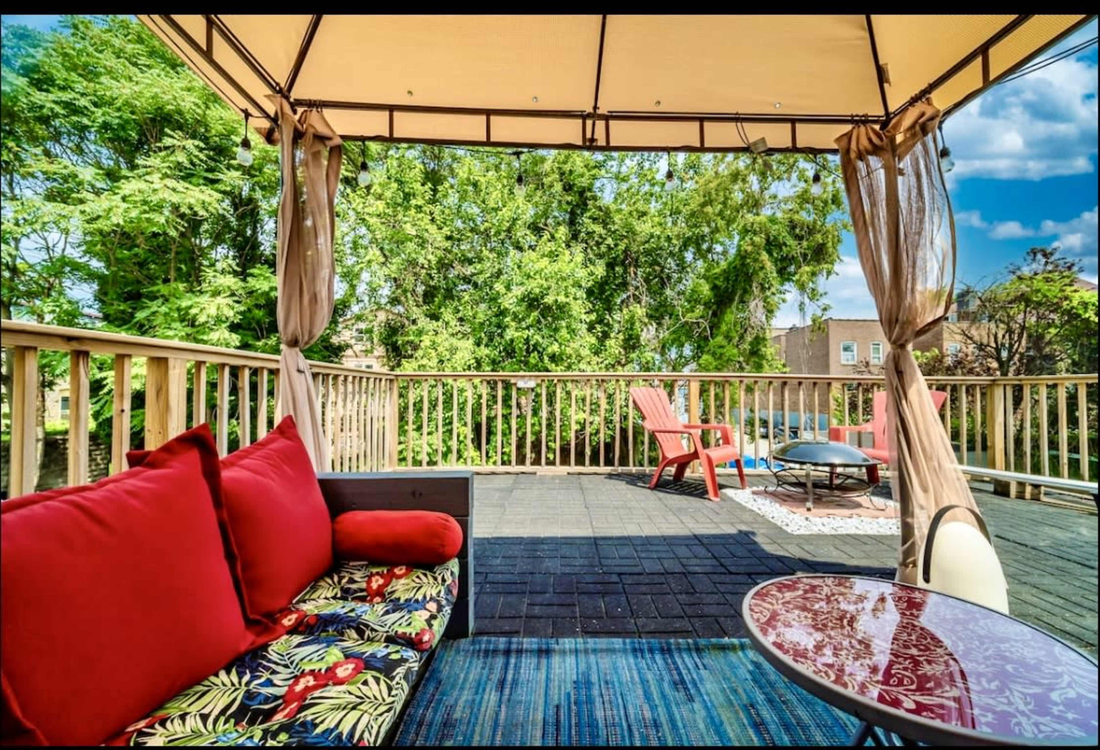 A patio featuring a gazebo with draped curtains, colorful seating options, and a view of trees.