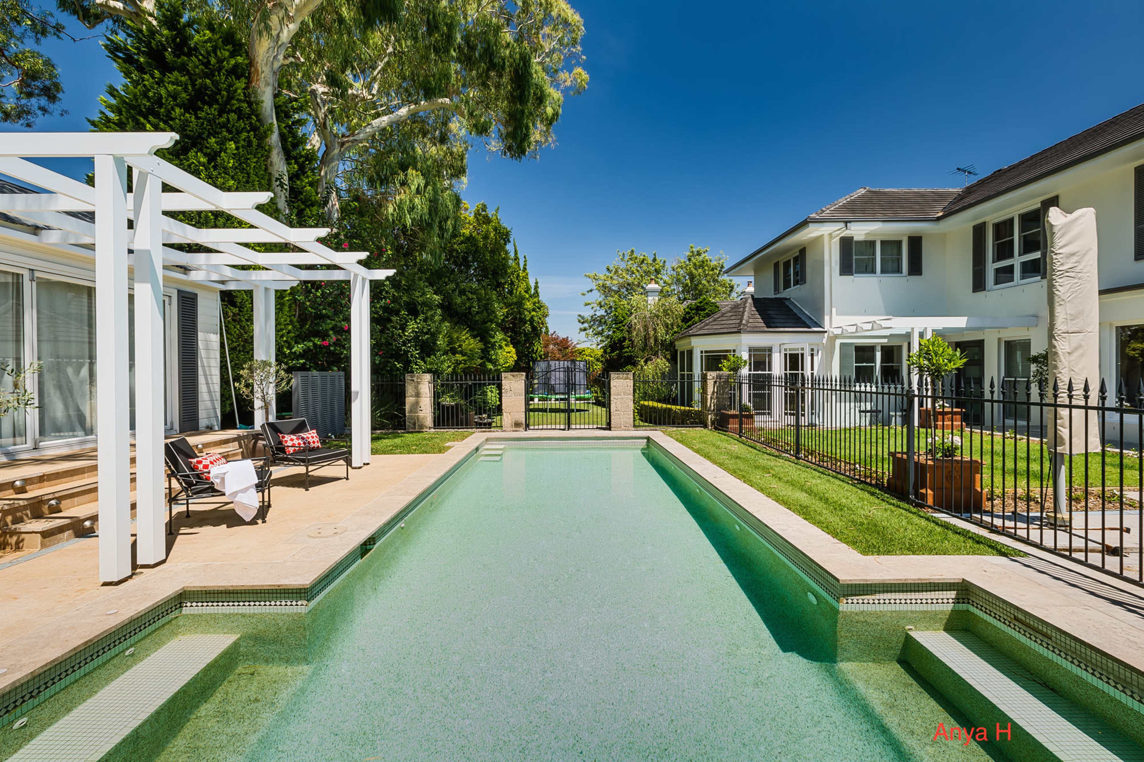 A clear swimming pool surrounded by a well-maintained garden and a two-story house in the background.