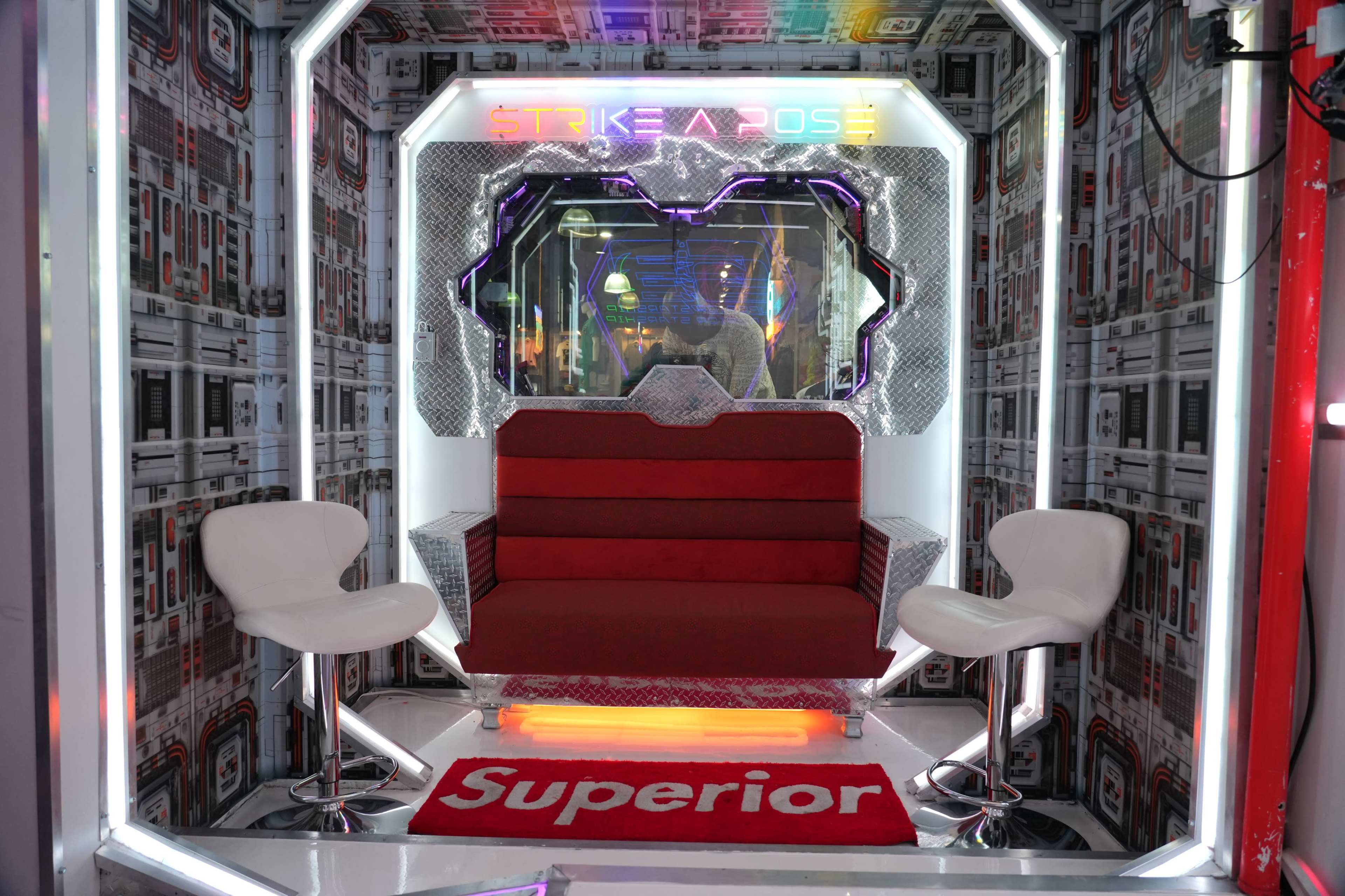A futuristic photo booth setup with a red sofa and two white chairs surrounded by patterned walls and colorful lighting.
