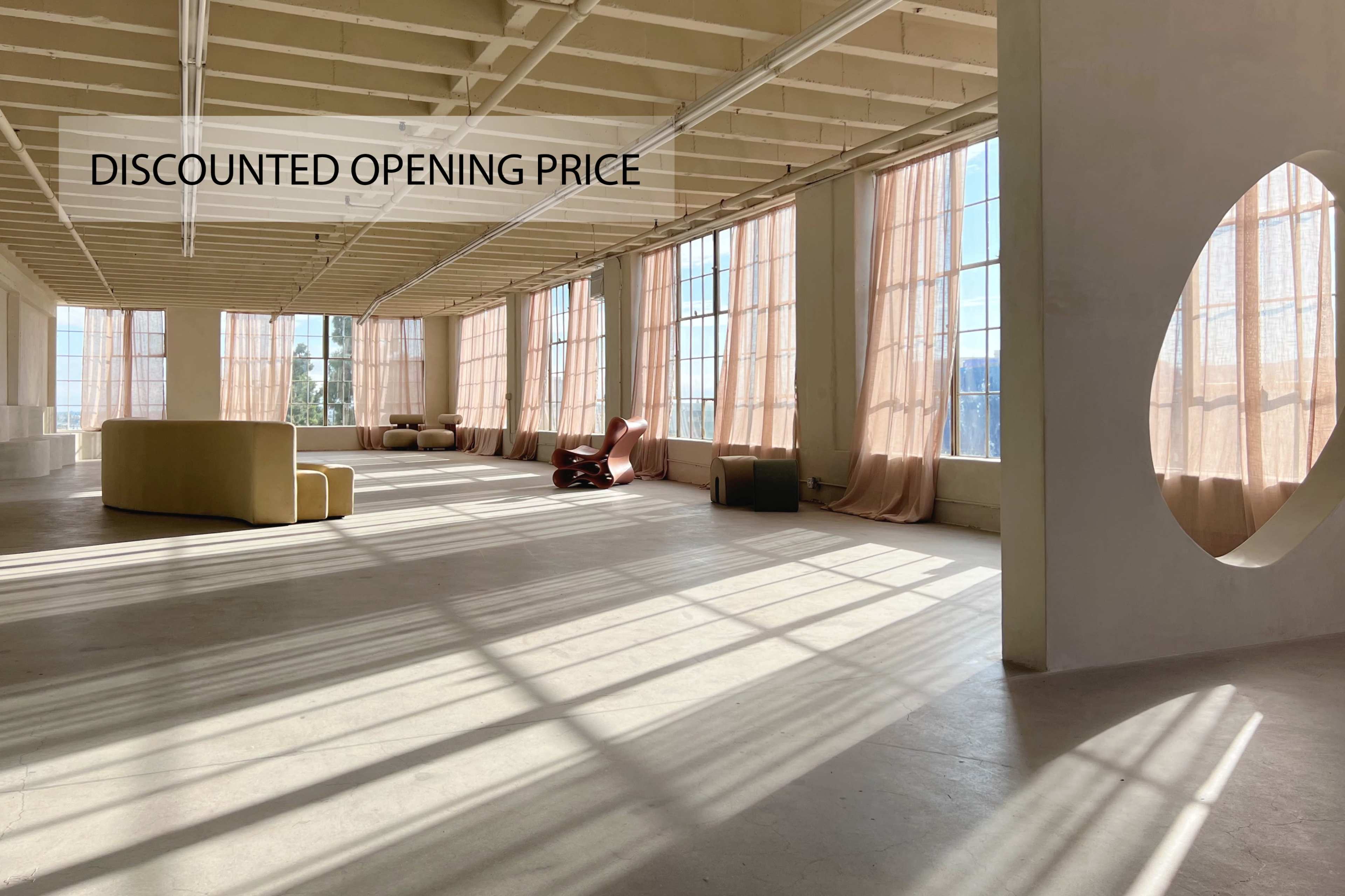 A spacious, well-lit room features large windows with sheer curtains, showcasing minimalist furniture and light streaming across the floor.