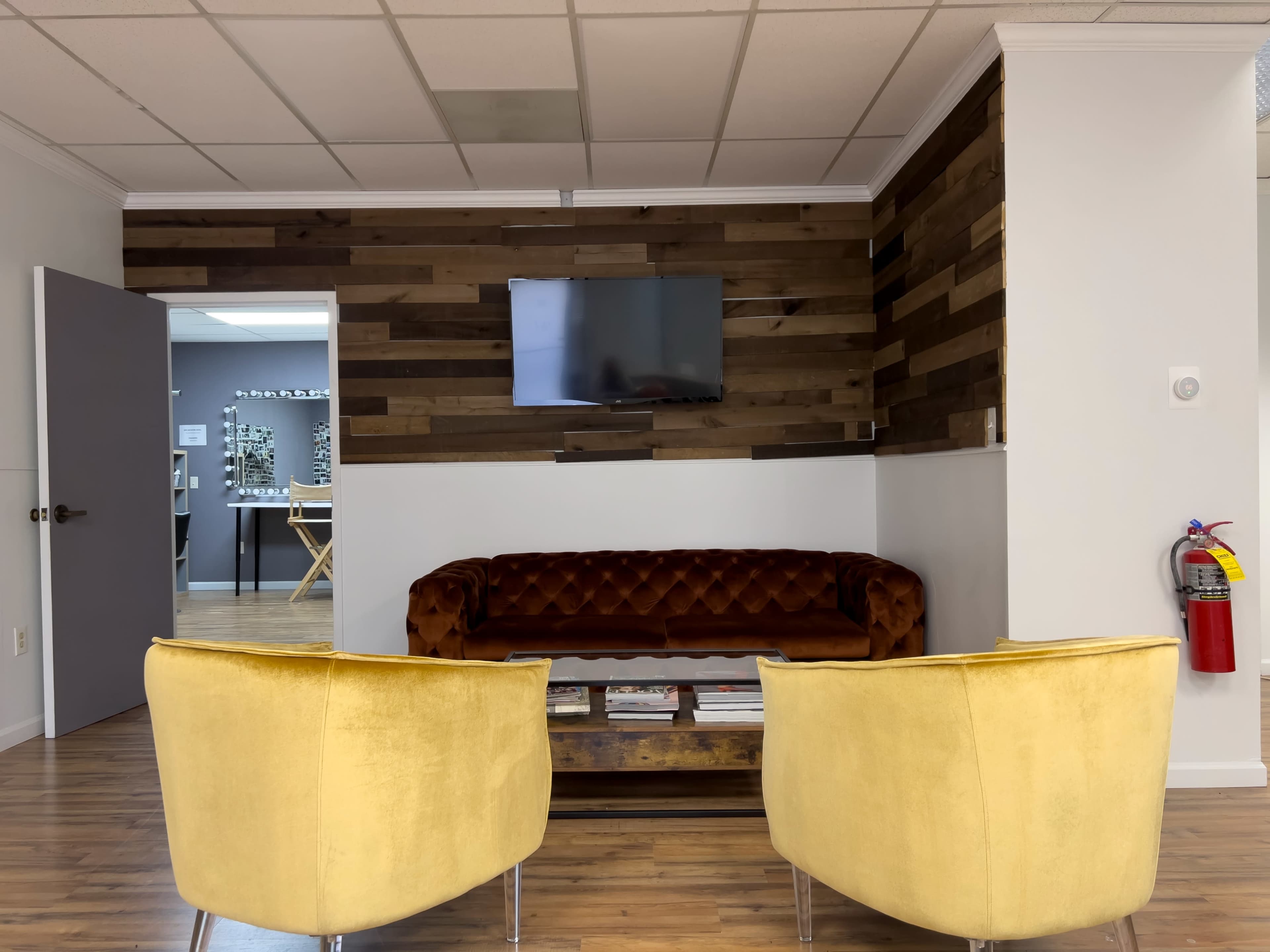 A waiting area with a brown tufted couch, two yellow armchairs, a wooden accent wall, a flat-screen TV, and a fire extinguisher mounted on the wall.
