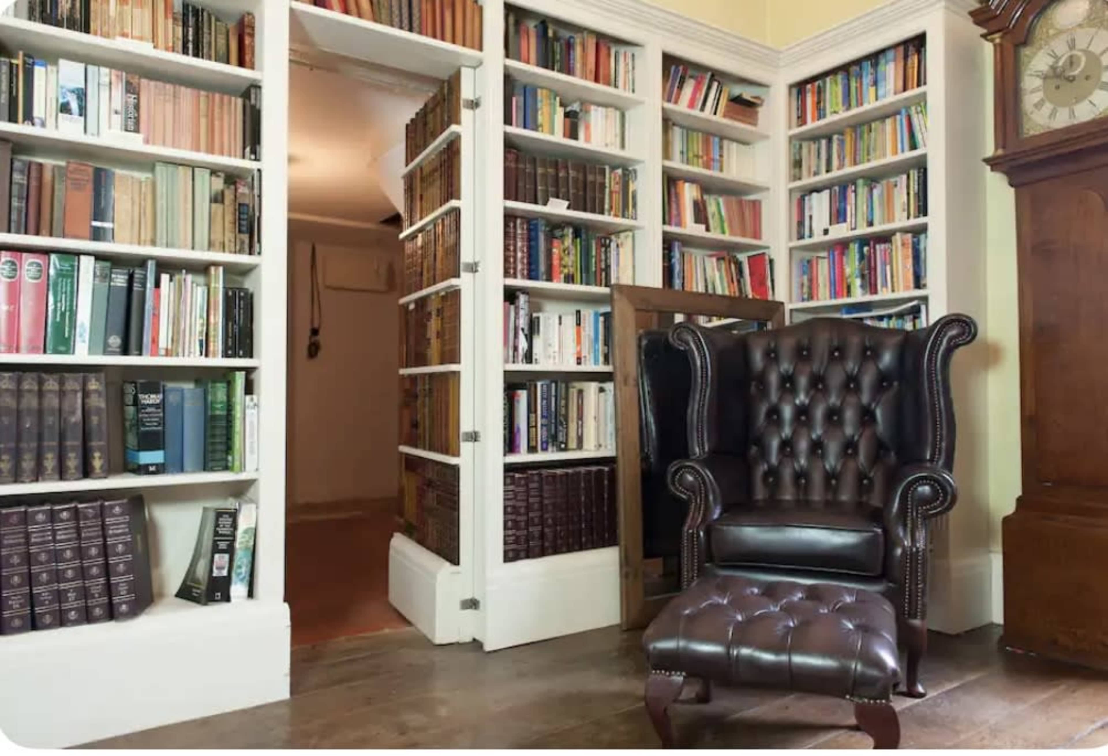 A leather armchair sits in a room with white bookshelves filled with numerous books, and a wooden grandfather clock is visible in the corner.