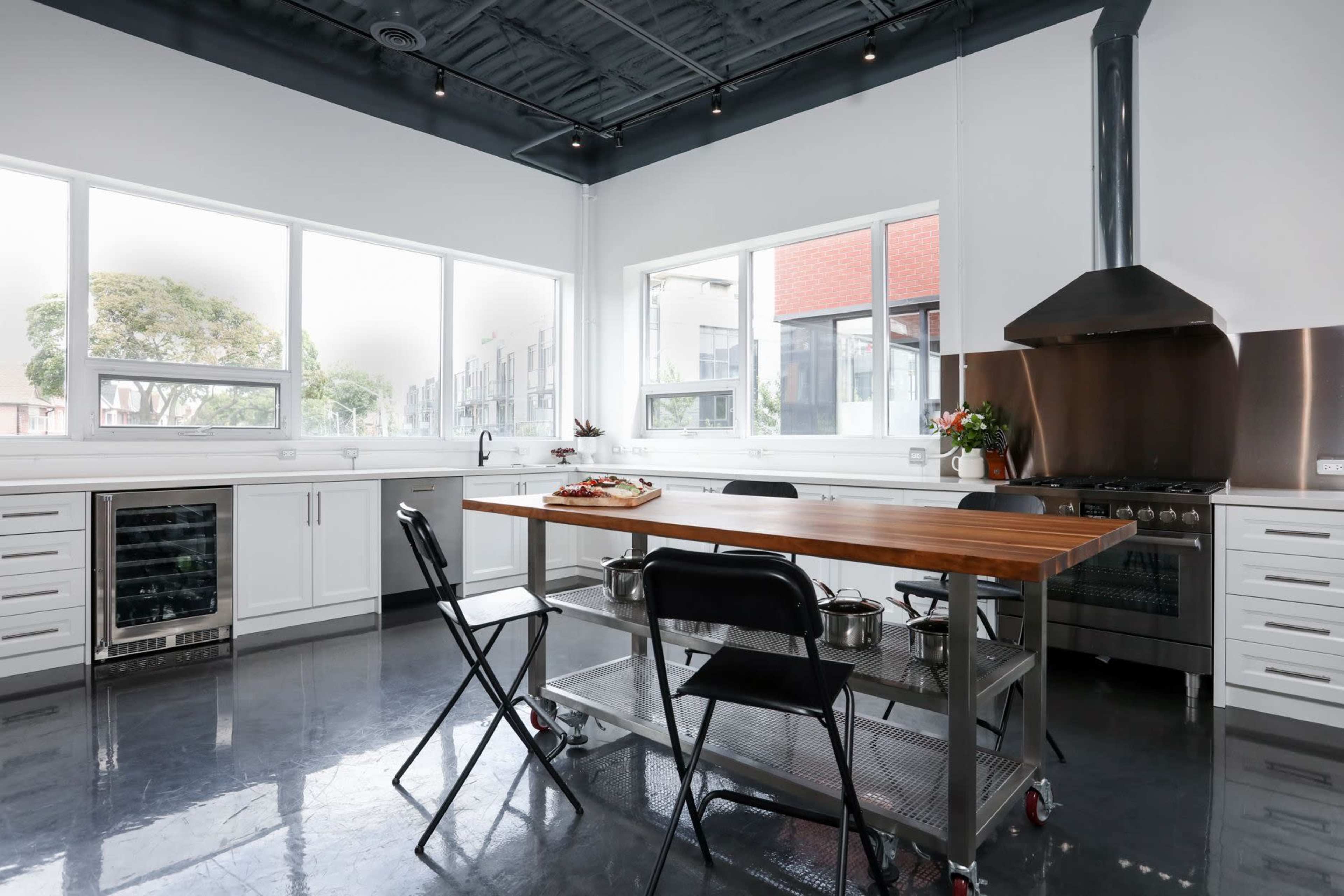 A modern kitchen features a central wooden table surrounded by black chairs, stainless steel appliances, and large windows that allow natural light to enter.