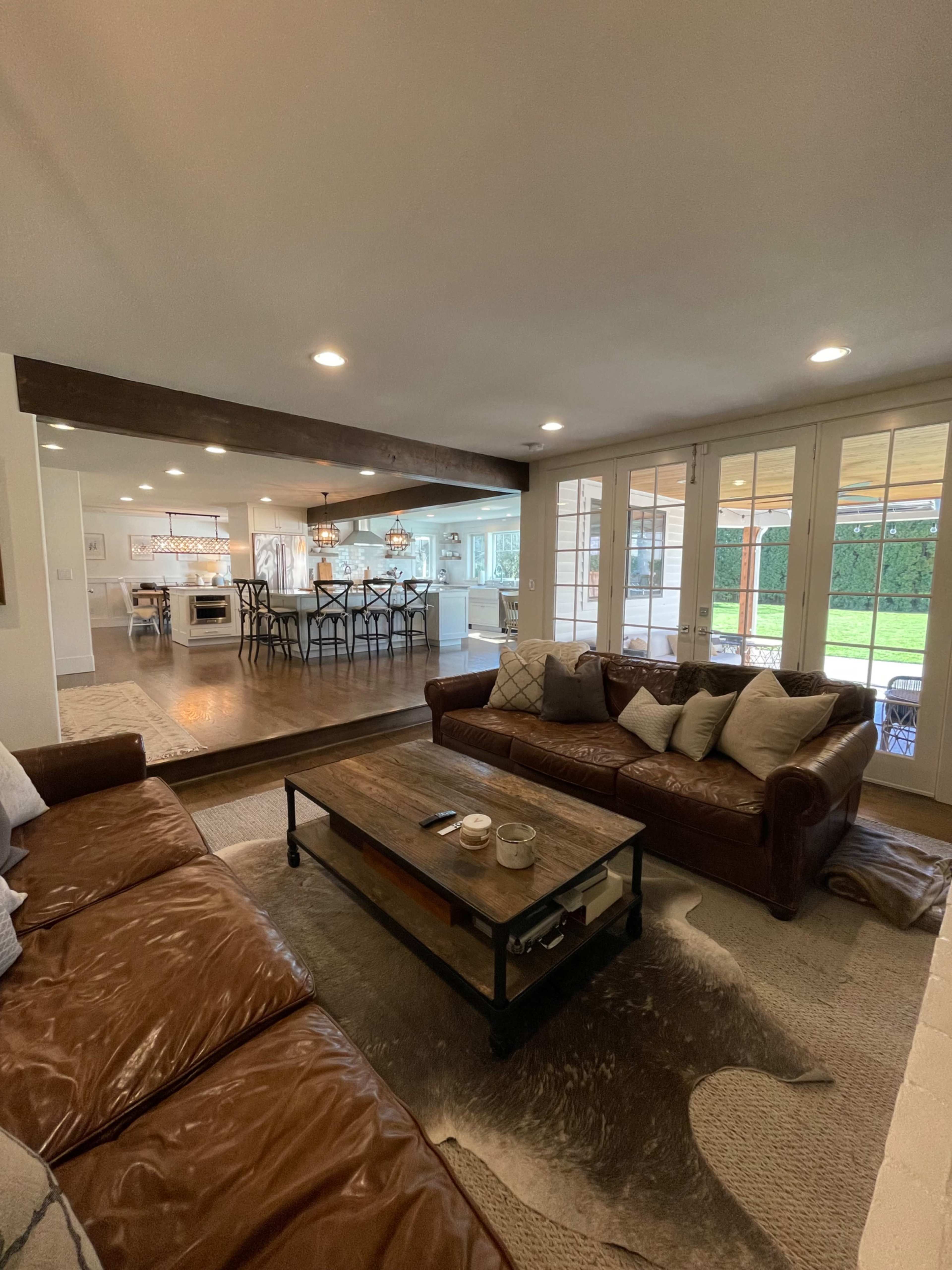 A cozy living room features two leather sofas facing a wooden coffee table, with large glass doors leading to a patio and an open kitchen in the background.